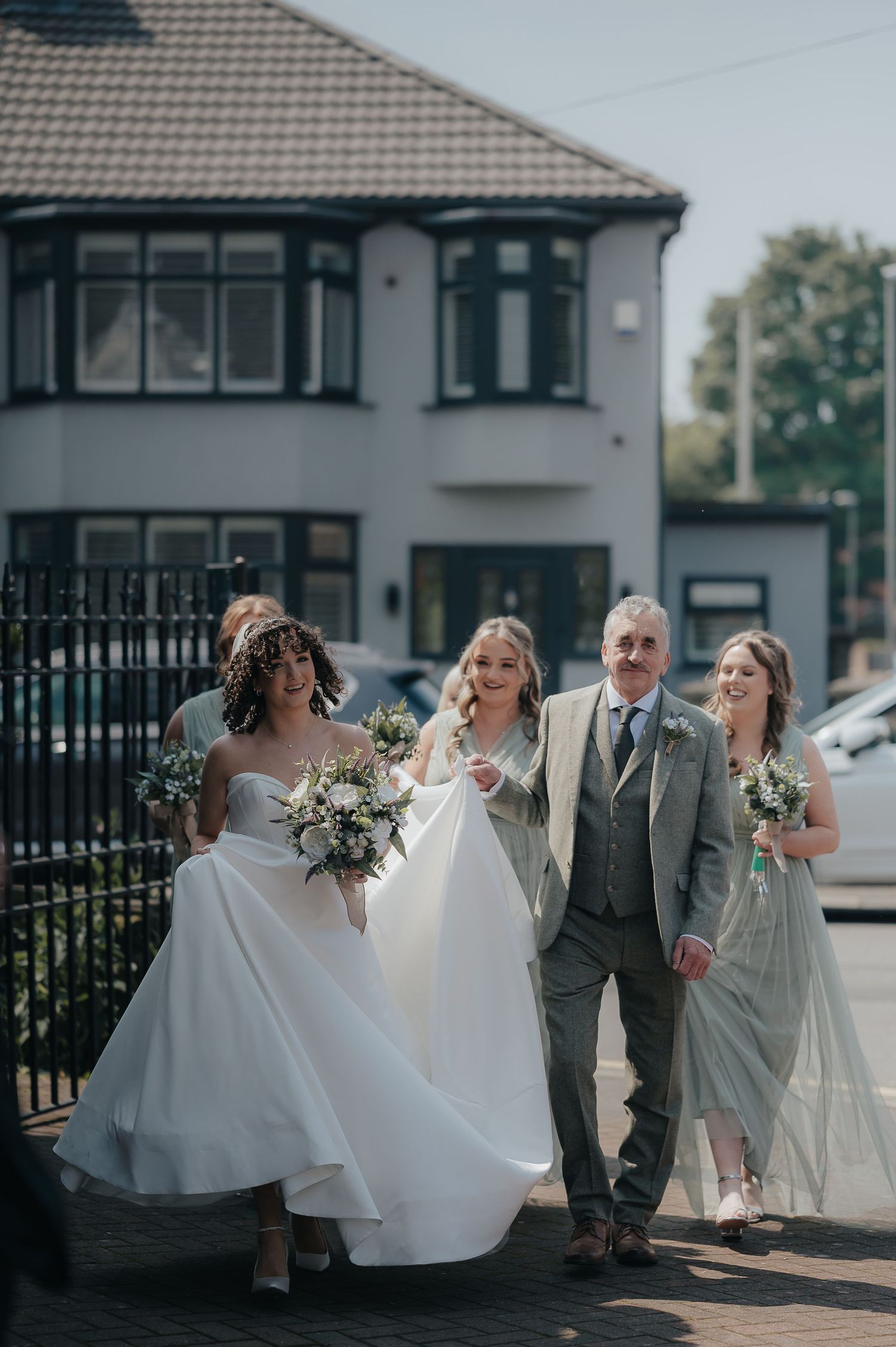 A wedding party walking outside a suburban house, with flowing dresses and bouquets.
