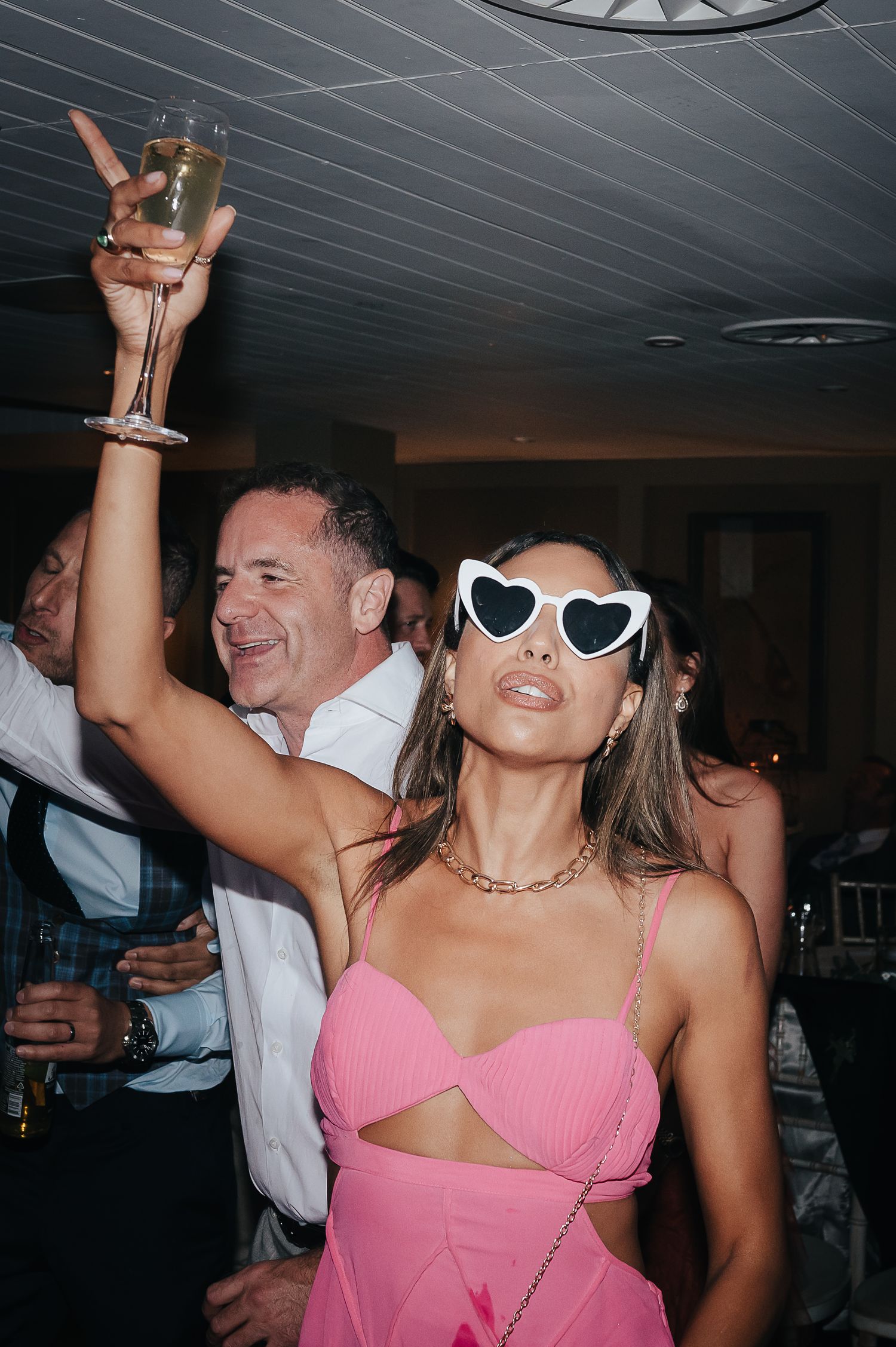 Party guest in pink dress and heart sunglasses raises champagne glass while dancing.