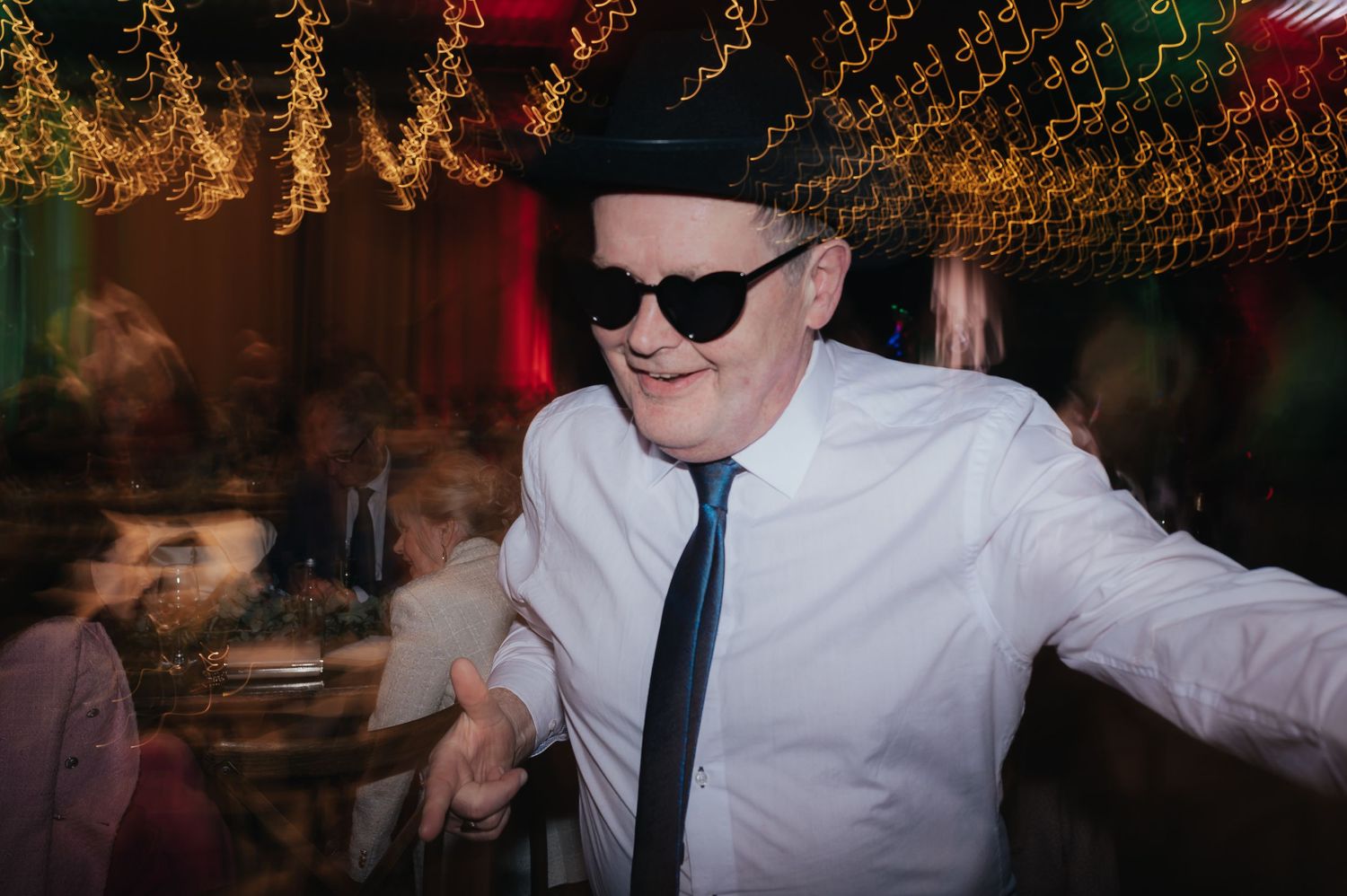 Wedding guest in sunglasses and tie dancing enthusiastically under warm lighting.