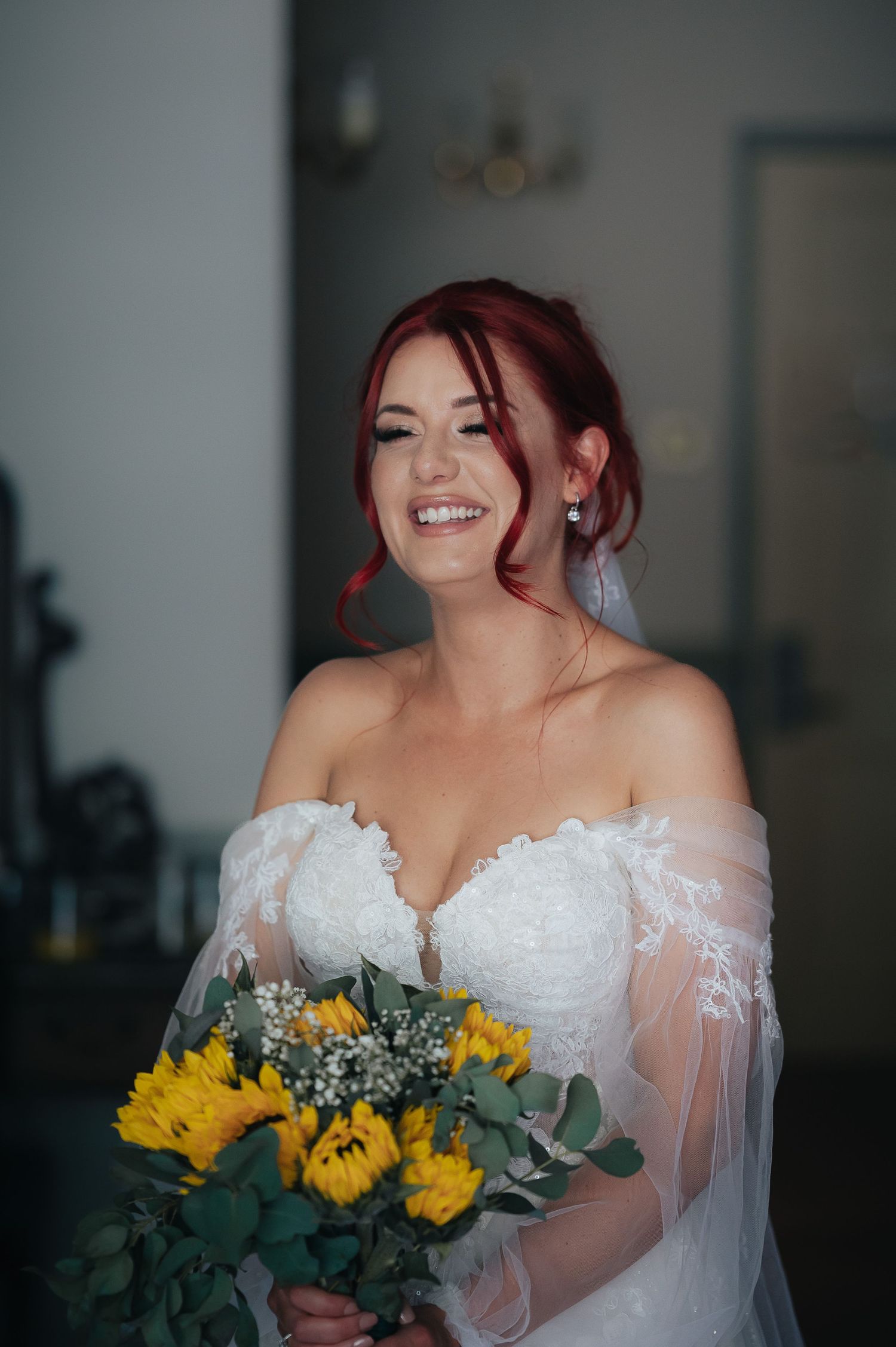 Bride wearing off-shoulder lace gown holds sunflower bouquet while smiling radiantly.