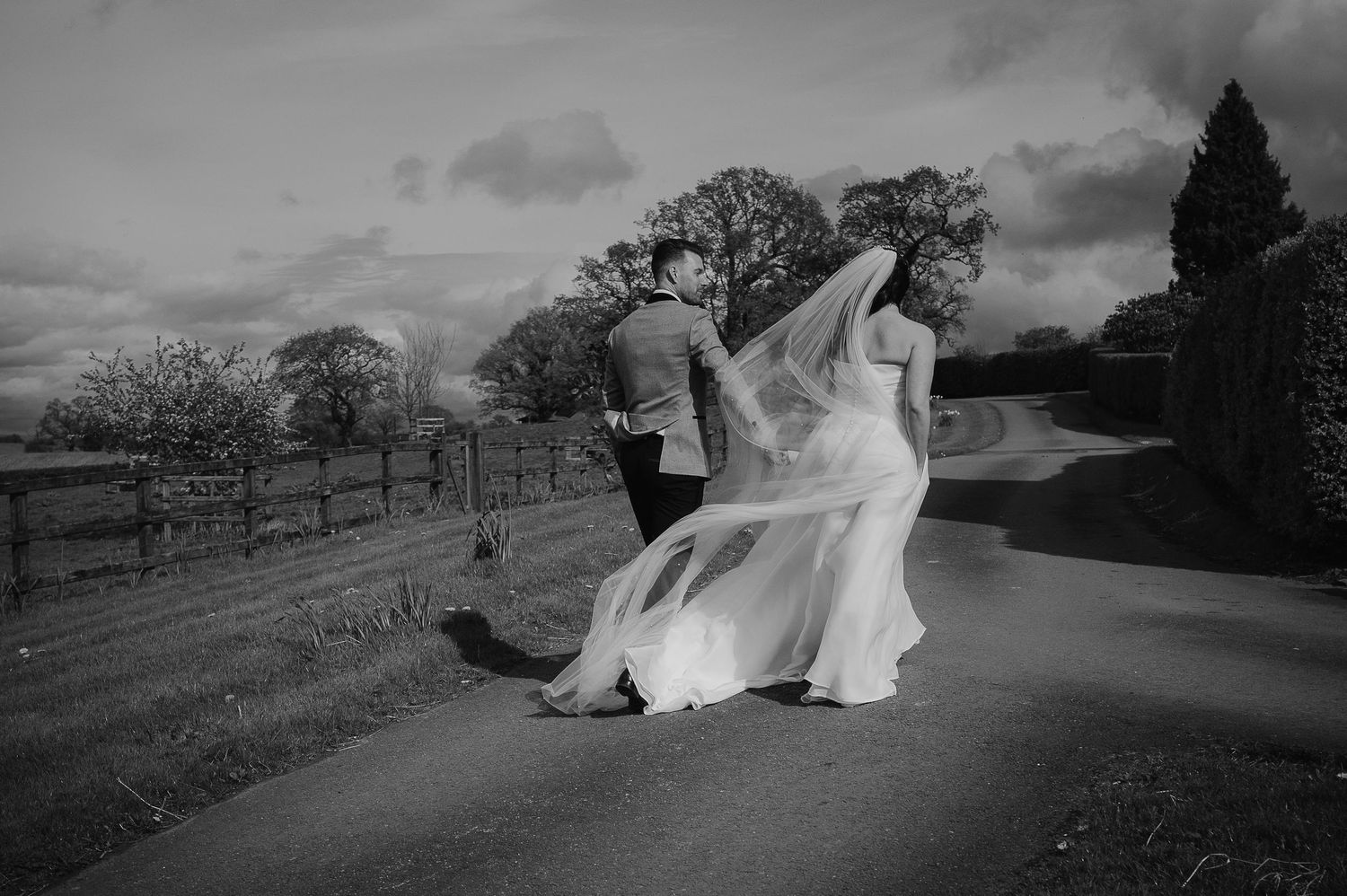 Black and white photo of bride's veil flowing dramatically in wind on country road.