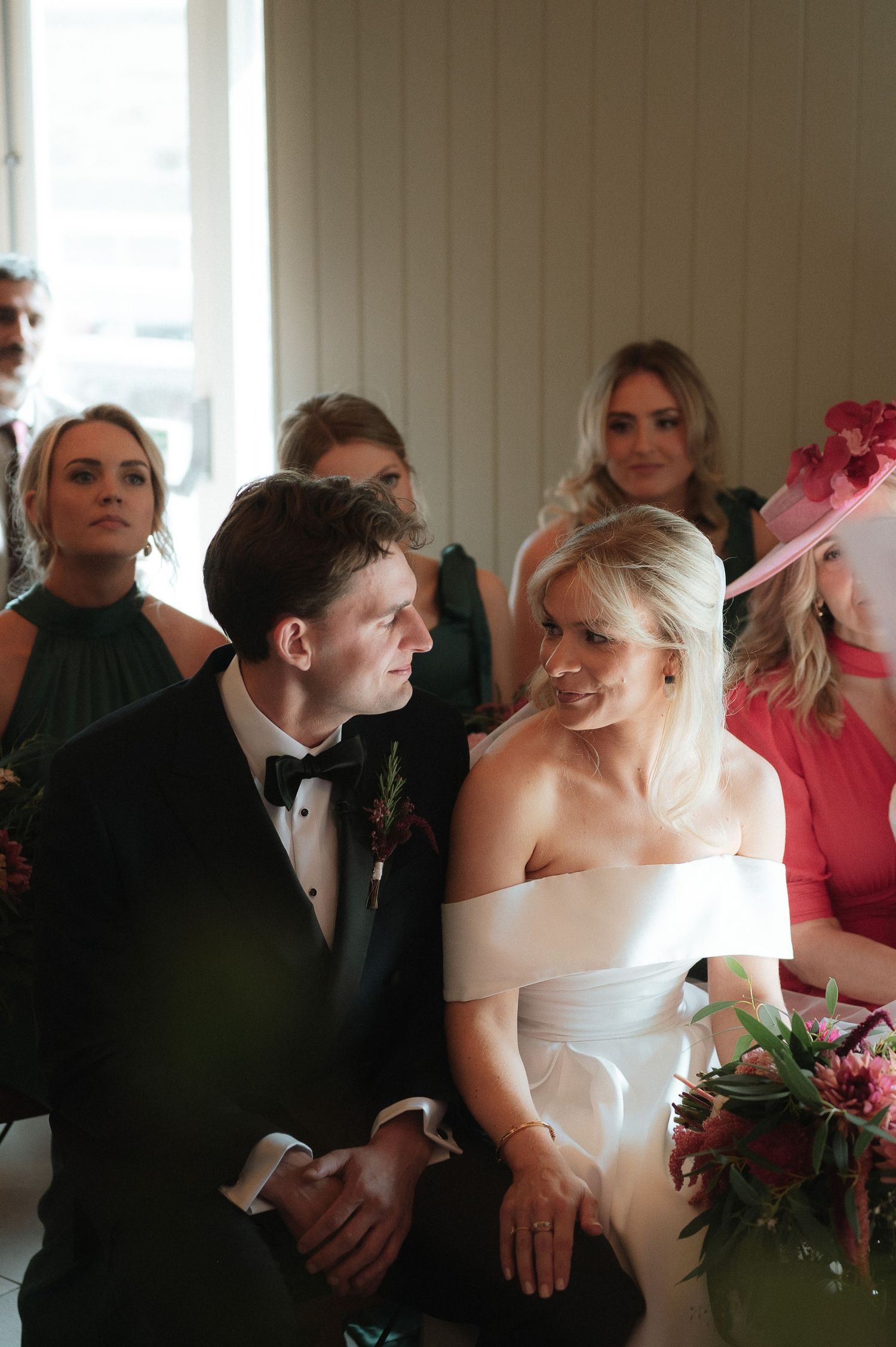 Wedding ceremony guests gather in an intimate indoor setting with white decor and floral arrangements.