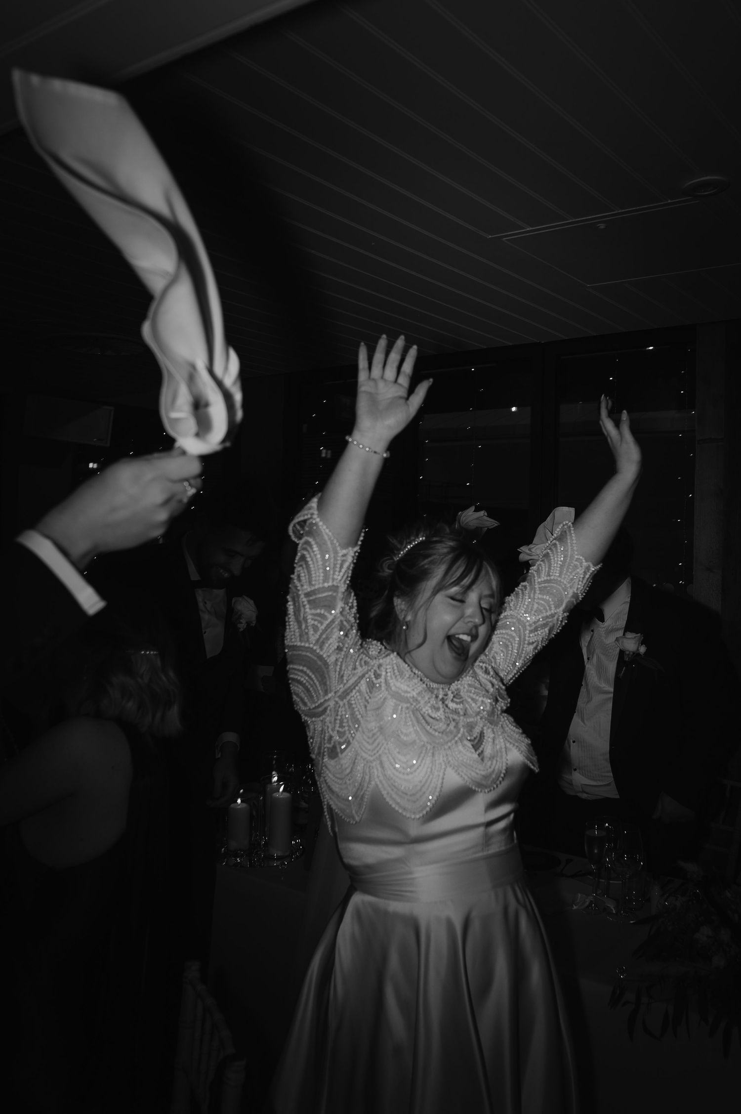 A person in a white wedding dress dances with arms raised during a celebration in a dark reception venue.