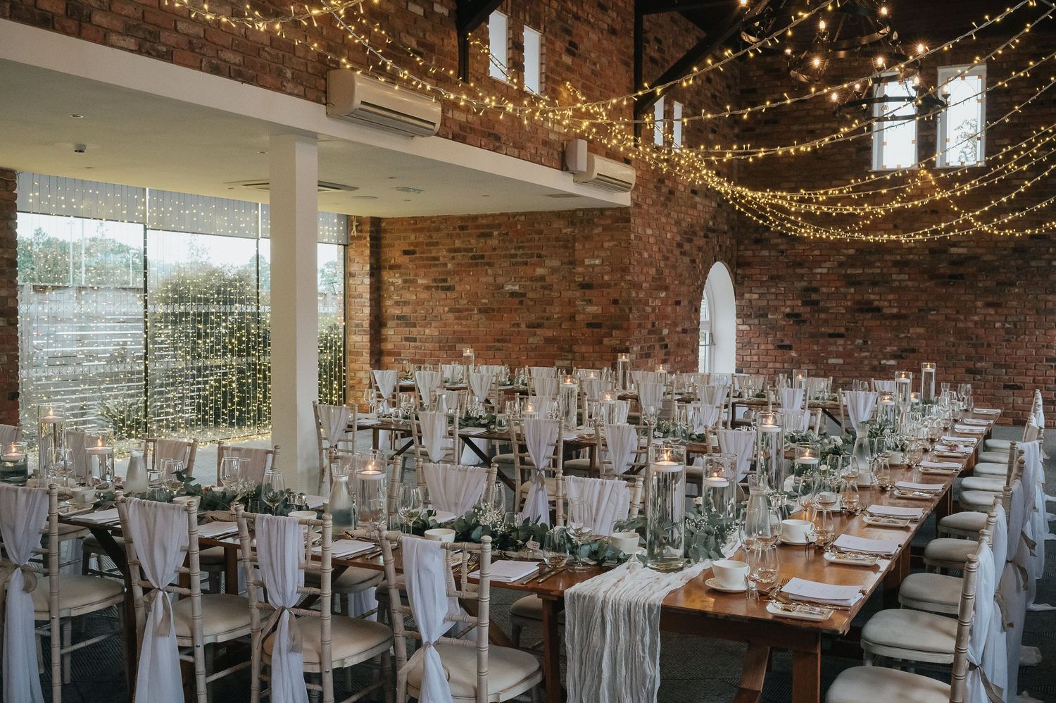 Wedding reception venue decorated with string lights and rustic brick walls featuring elegant table settings.