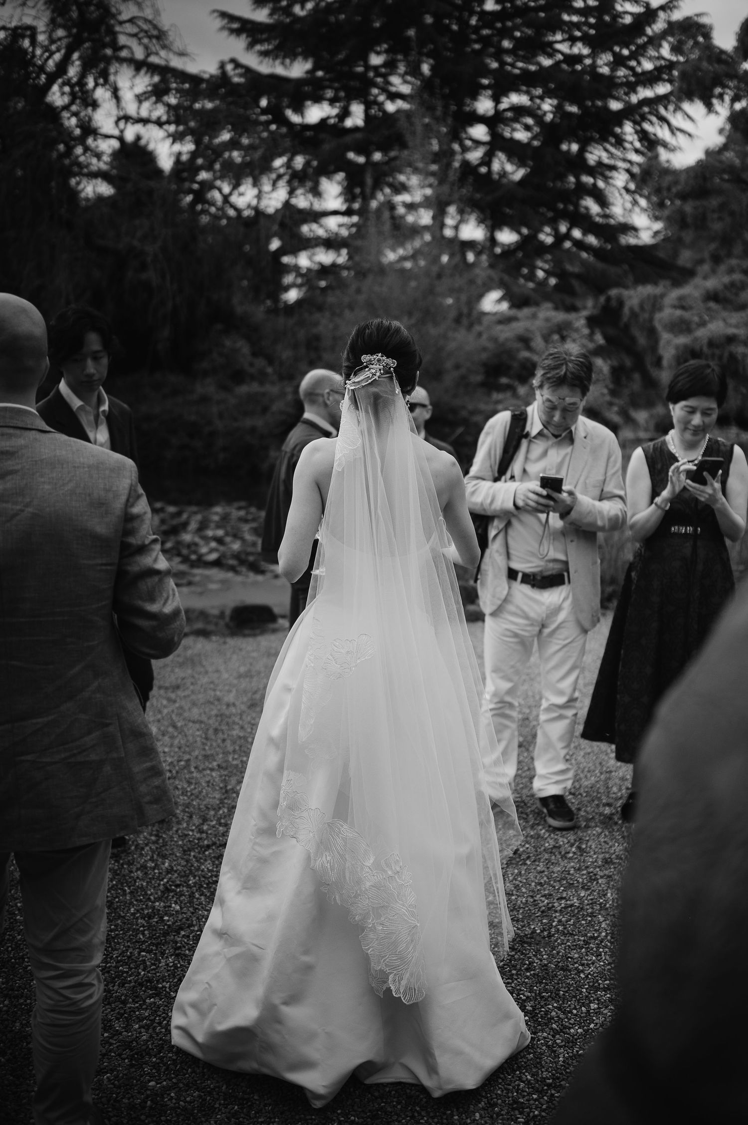 Bride in flowing wedding dress and veil stands among guests at outdoor garden ceremony.