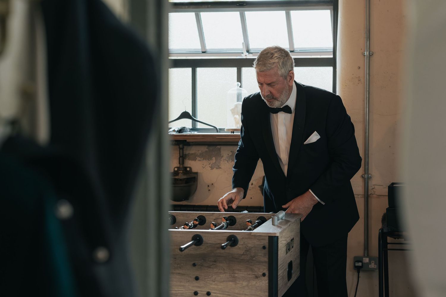 A person in a formal black tuxedo prepares in a room with natural light coming through the window.