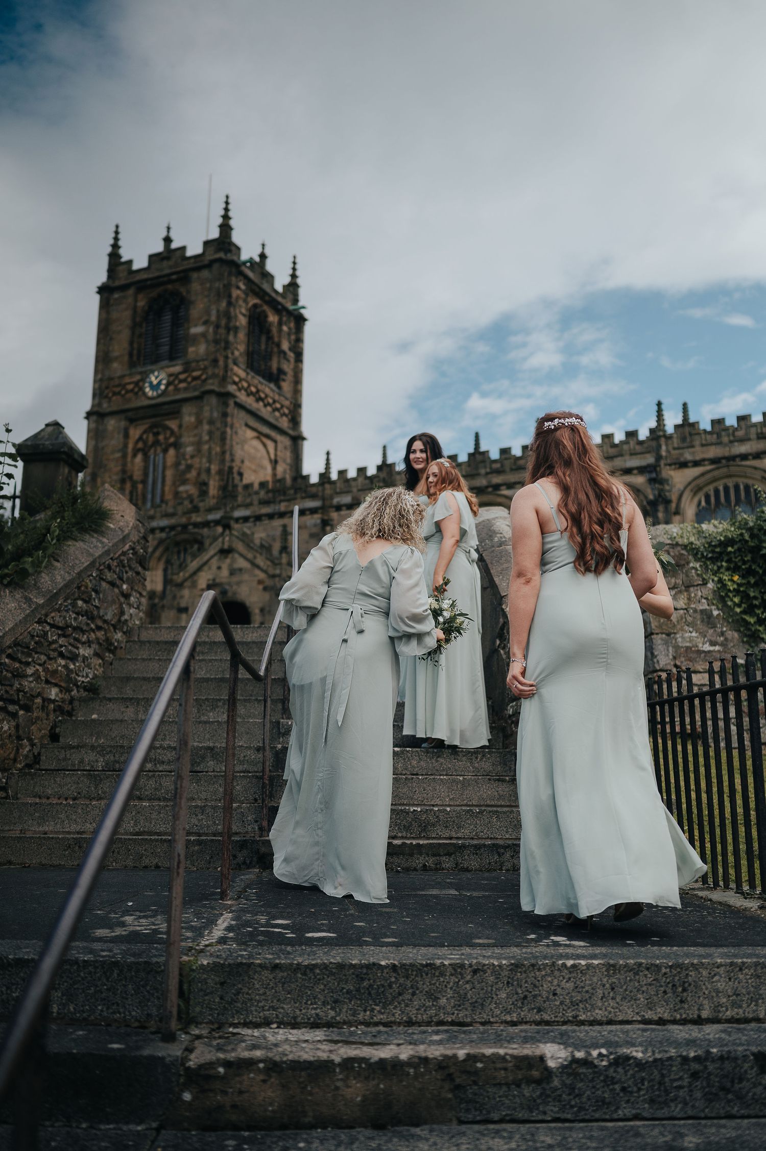 Bridesmaids in pale blue dresses ascending stone steps of a historic church with gothic architecture.