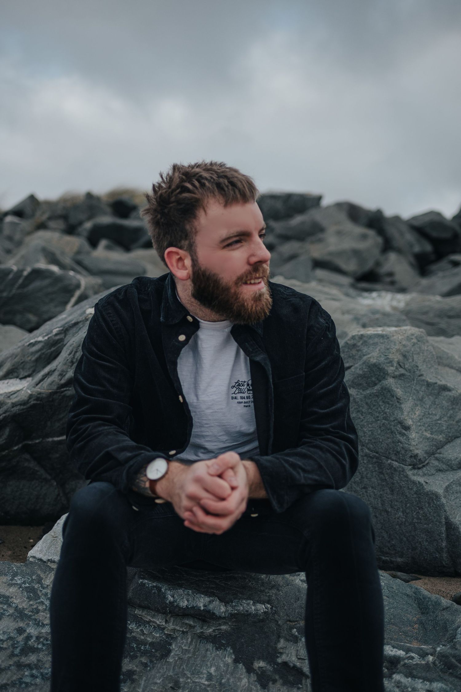 Person in black jacket sits thoughtfully on rocky coastline against moody gray sky at sunset.