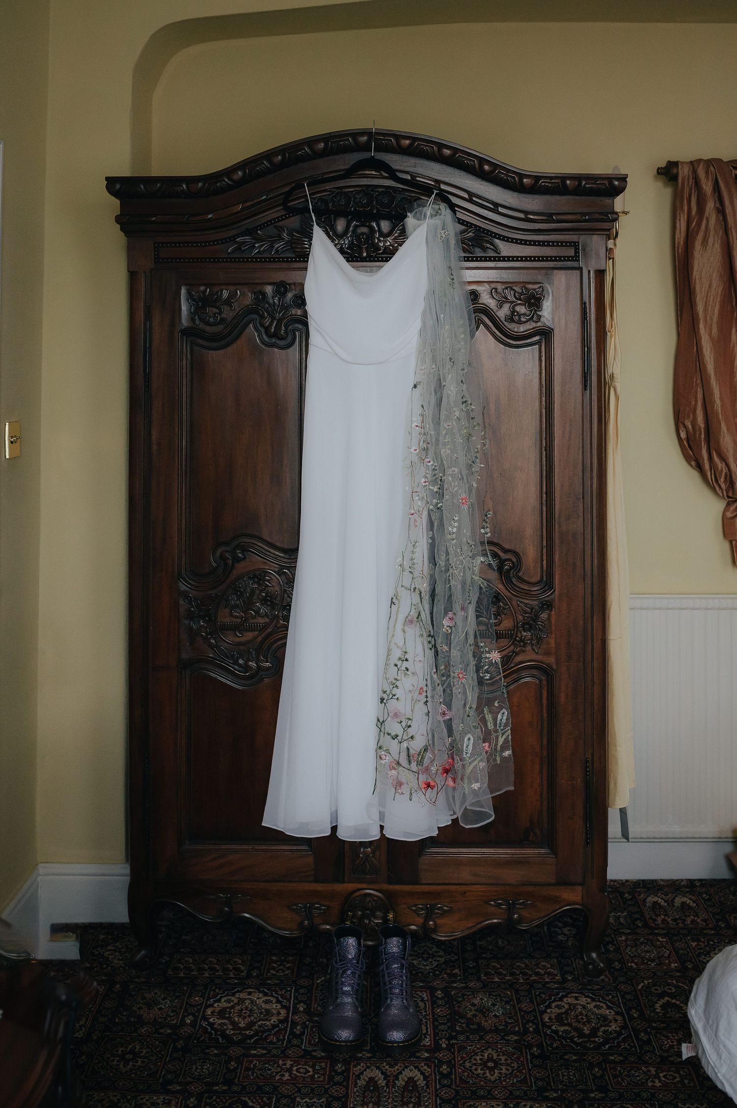 White wedding dress and veil hanging on ornate wooden wardrobe with shoes below.