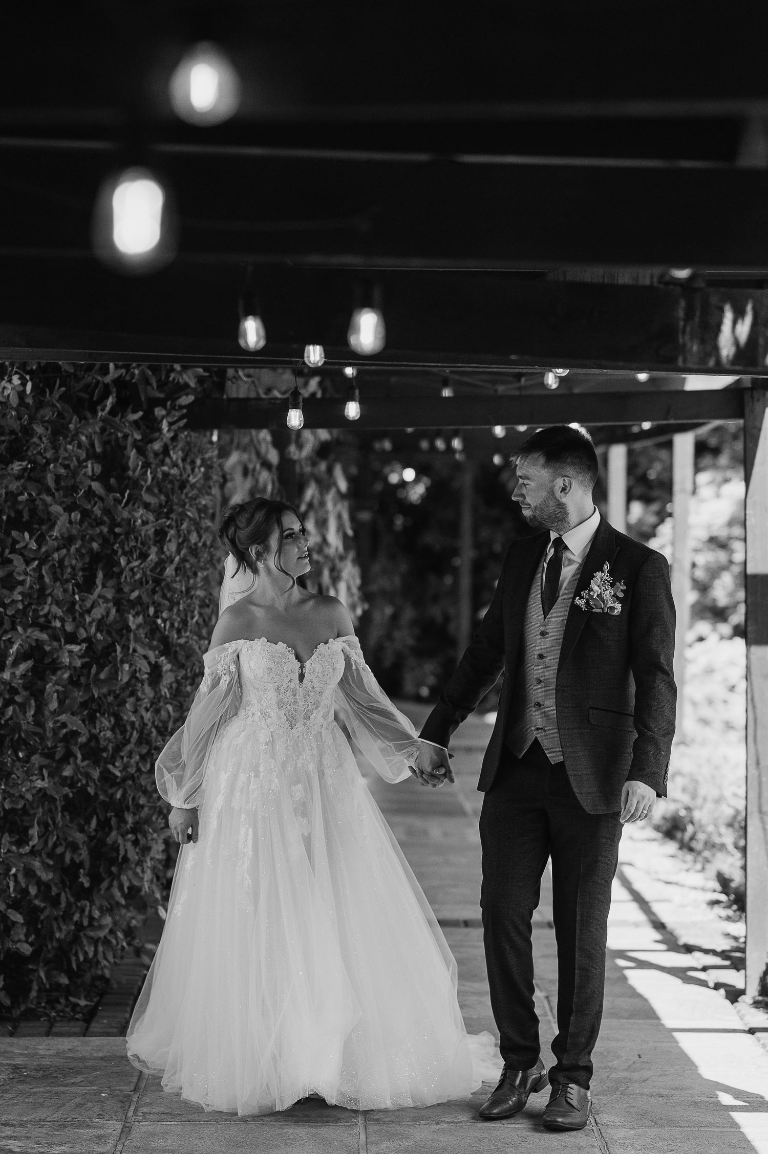 A black and white photo of a couple in wedding attire holding hands under a covered walkway.