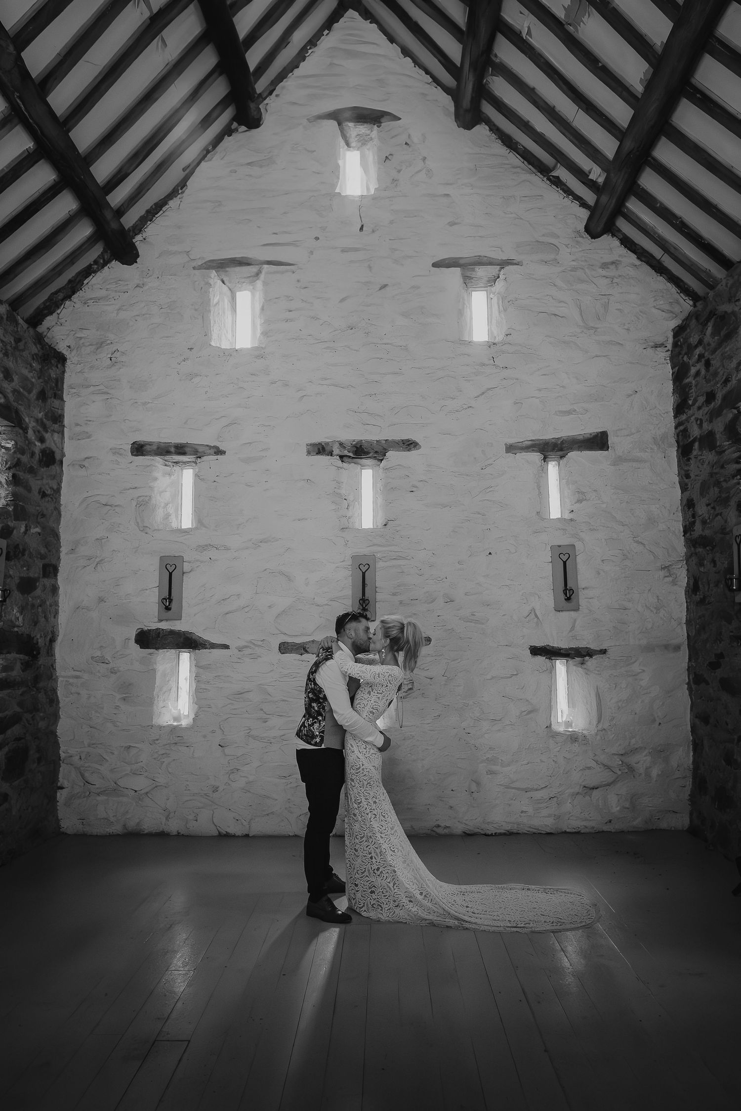 A couple embraces in front of a stone wall with nine illuminated windows in black and white.