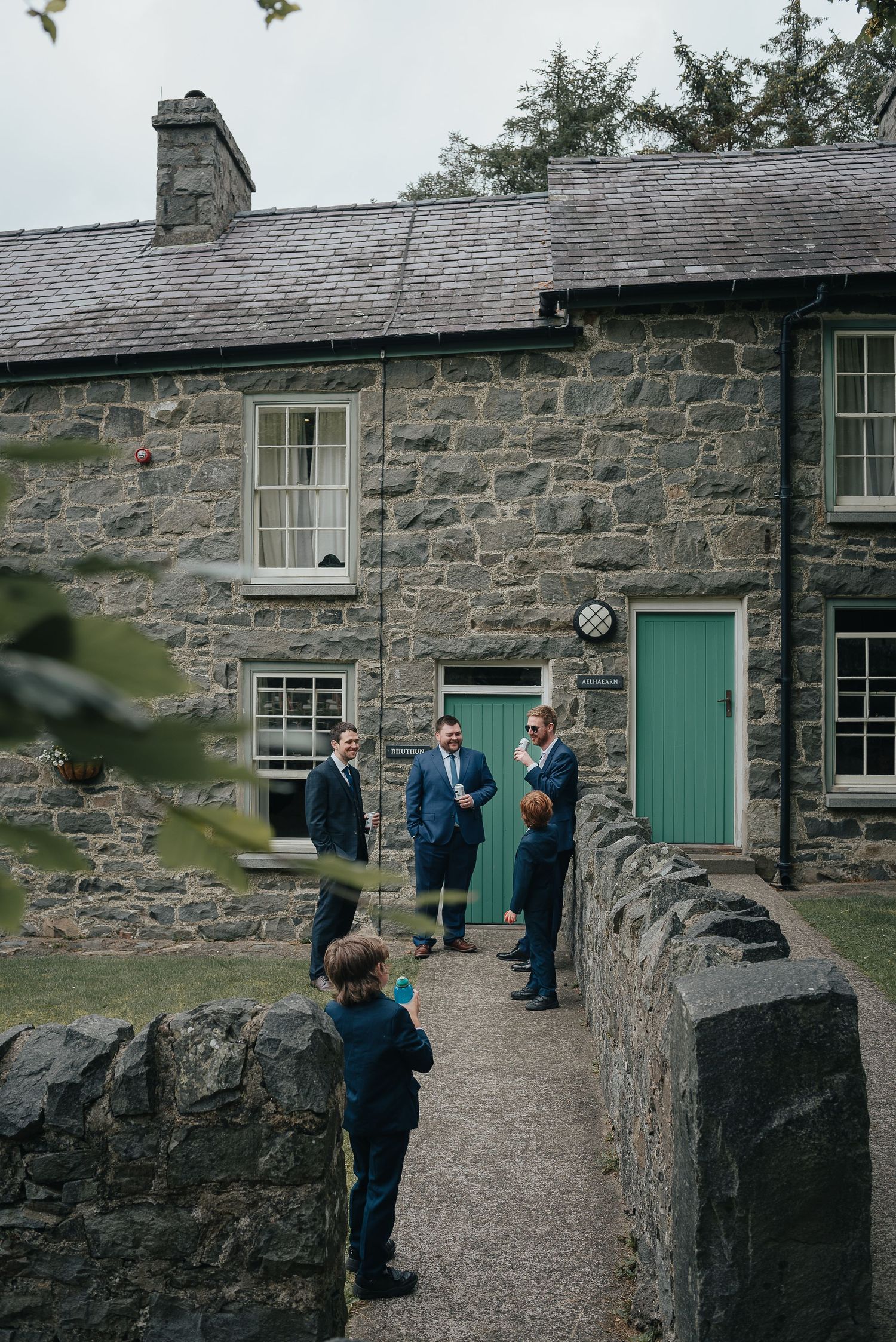 People gathered outside stone cottage with turquoise door during wedding preparations.