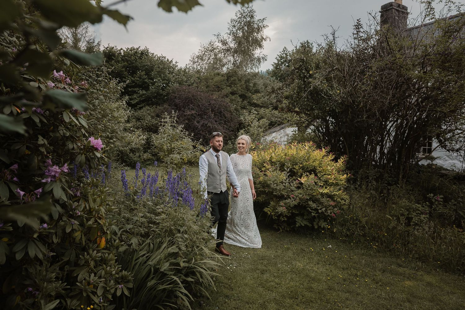 A couple strolls through a lush garden path with blooming flowers.