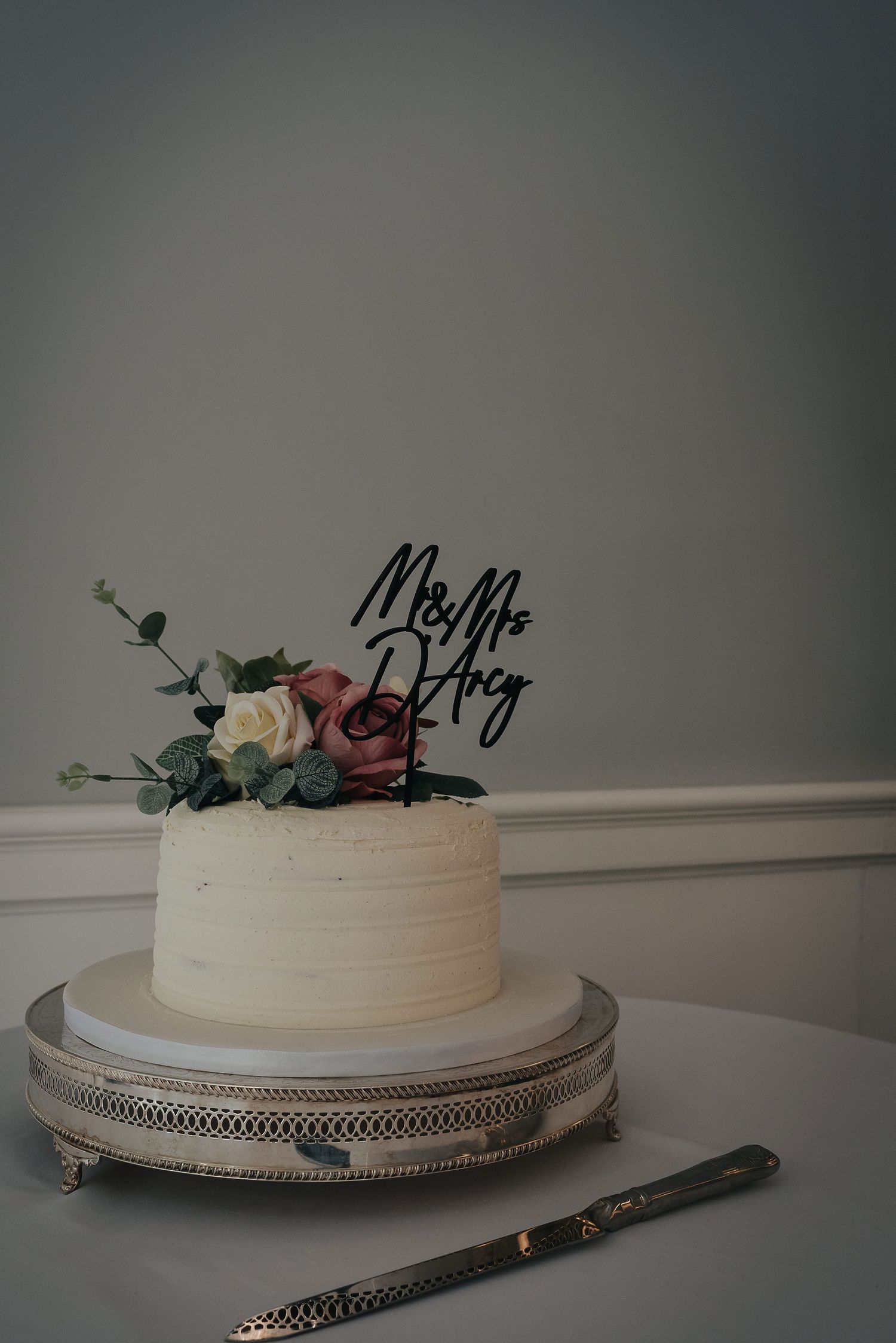 Simple white wedding cake with floral decorations and black 'Mr & Mrs' topper on silver stand.