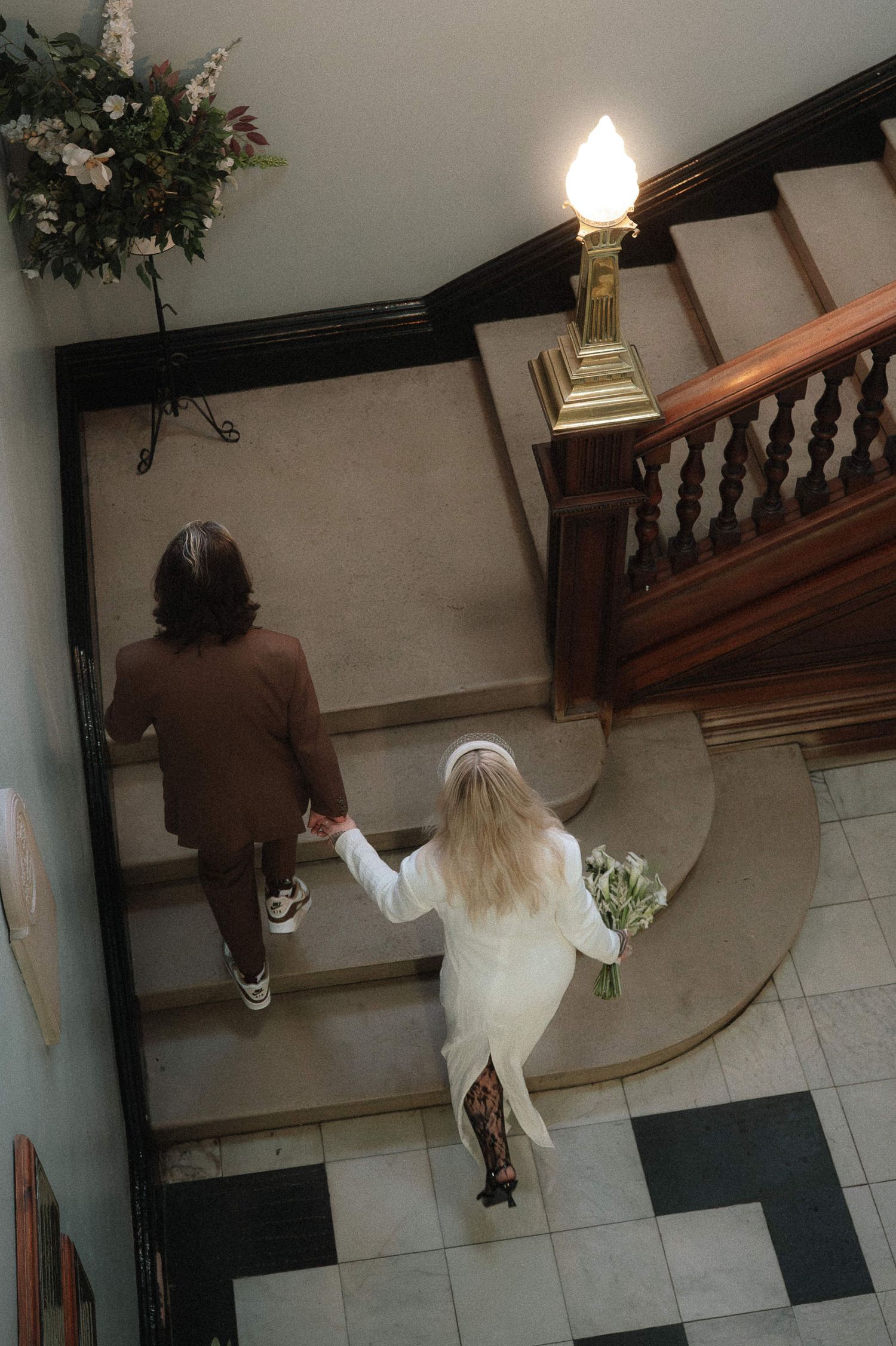 Two people walking up an elegant staircase with wooden railings and a decorative wall lamp.