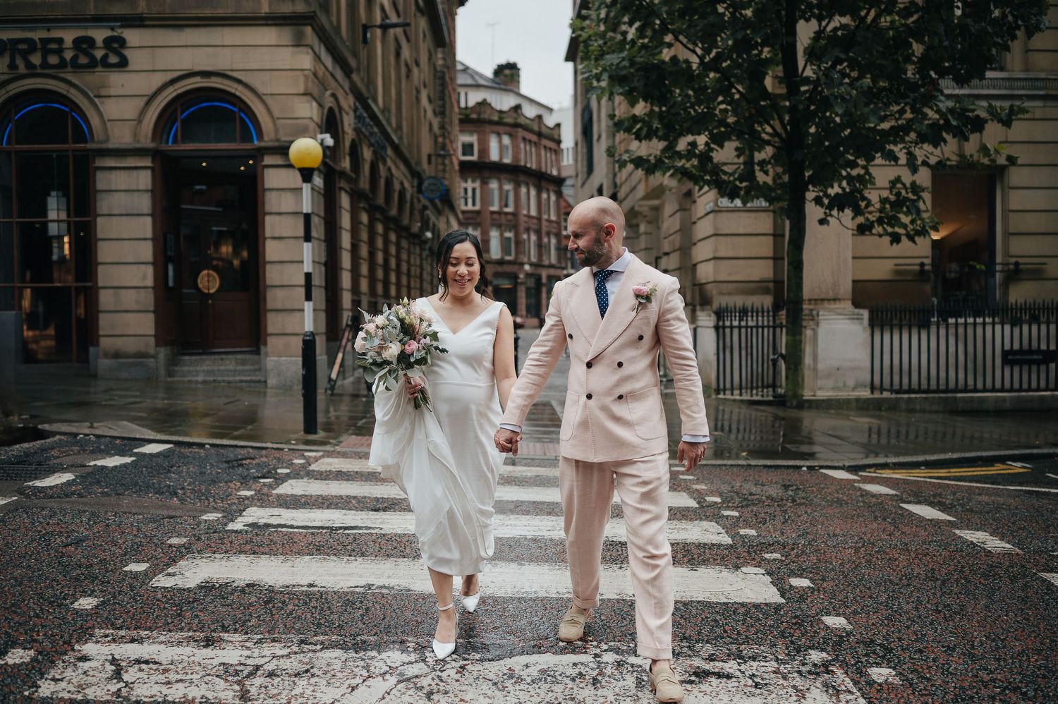 A wedding couple walks joyfully across a crosswalk in formal attire.