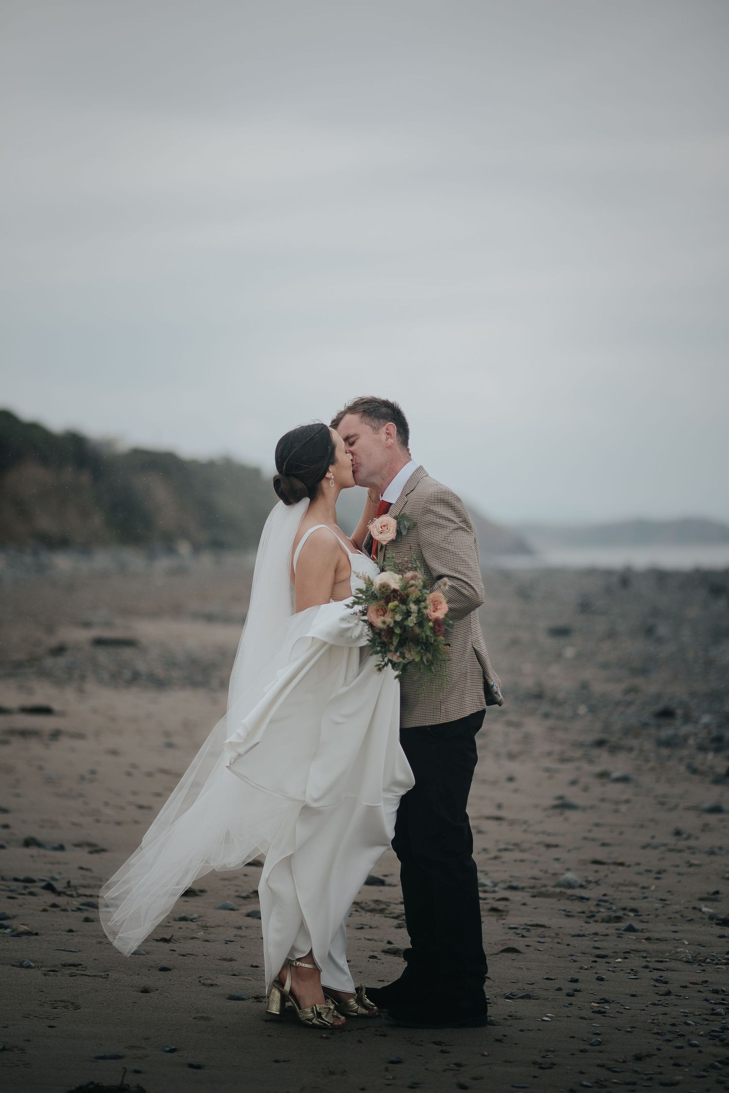 A romantic wedding couple embraces on a moody beach with the bride holding a floral bouquet.