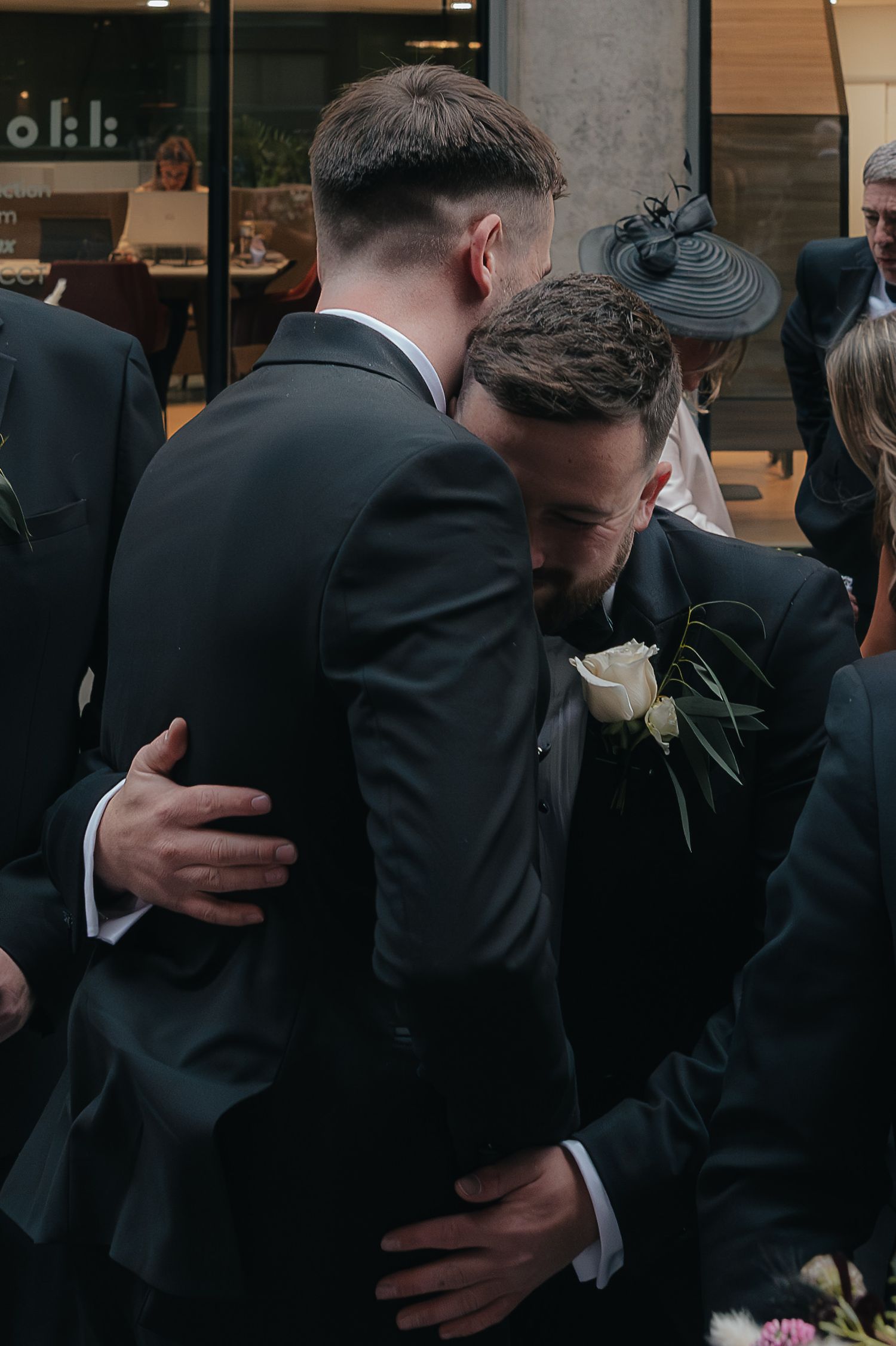 Two suited individuals embrace each other in a warm hug while standing at a formal event.