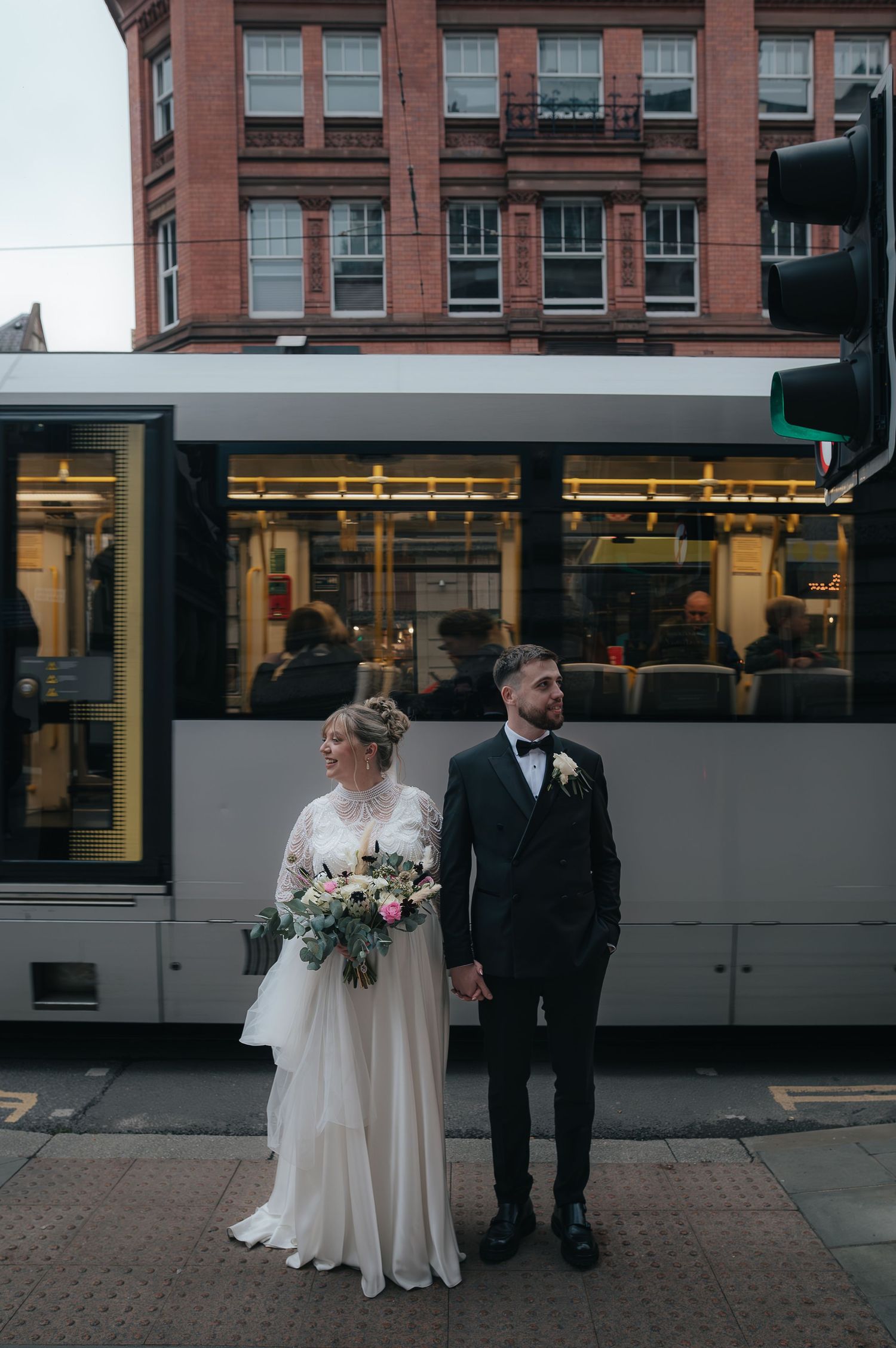 A couple in wedding attire stands on a city sidewalk as a streetcar passes behind them in front of a brick building.