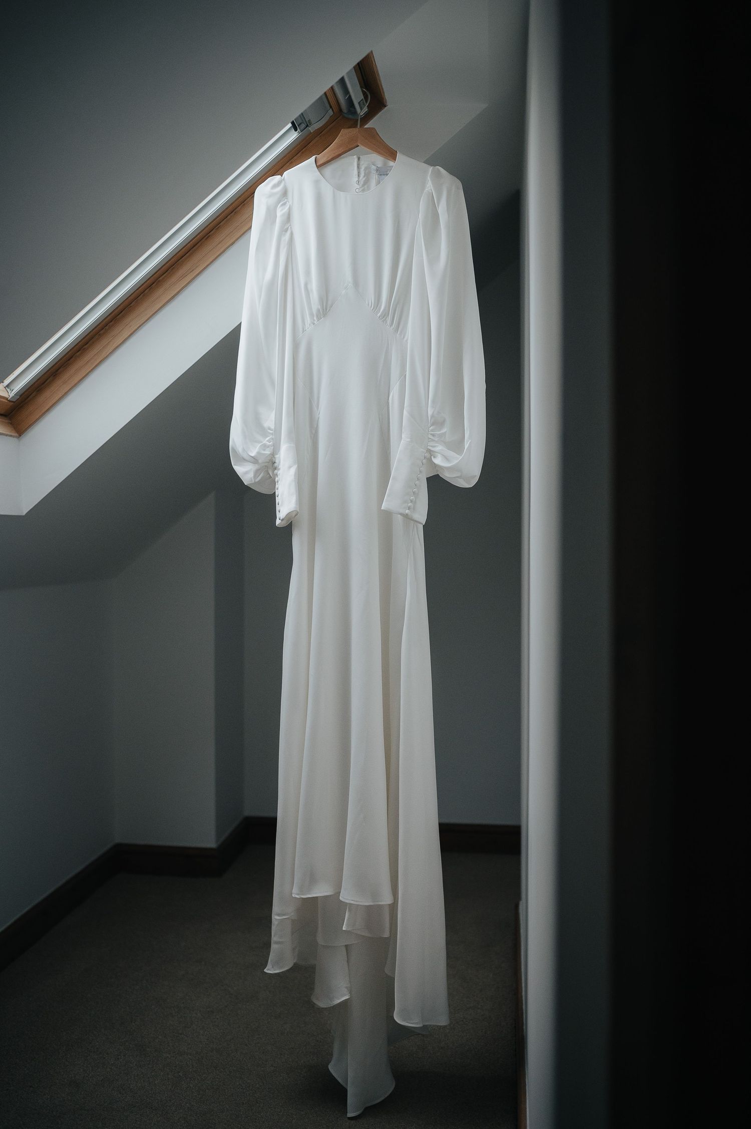 Long white wedding gown with flowing sleeves hanging in minimalist attic space.