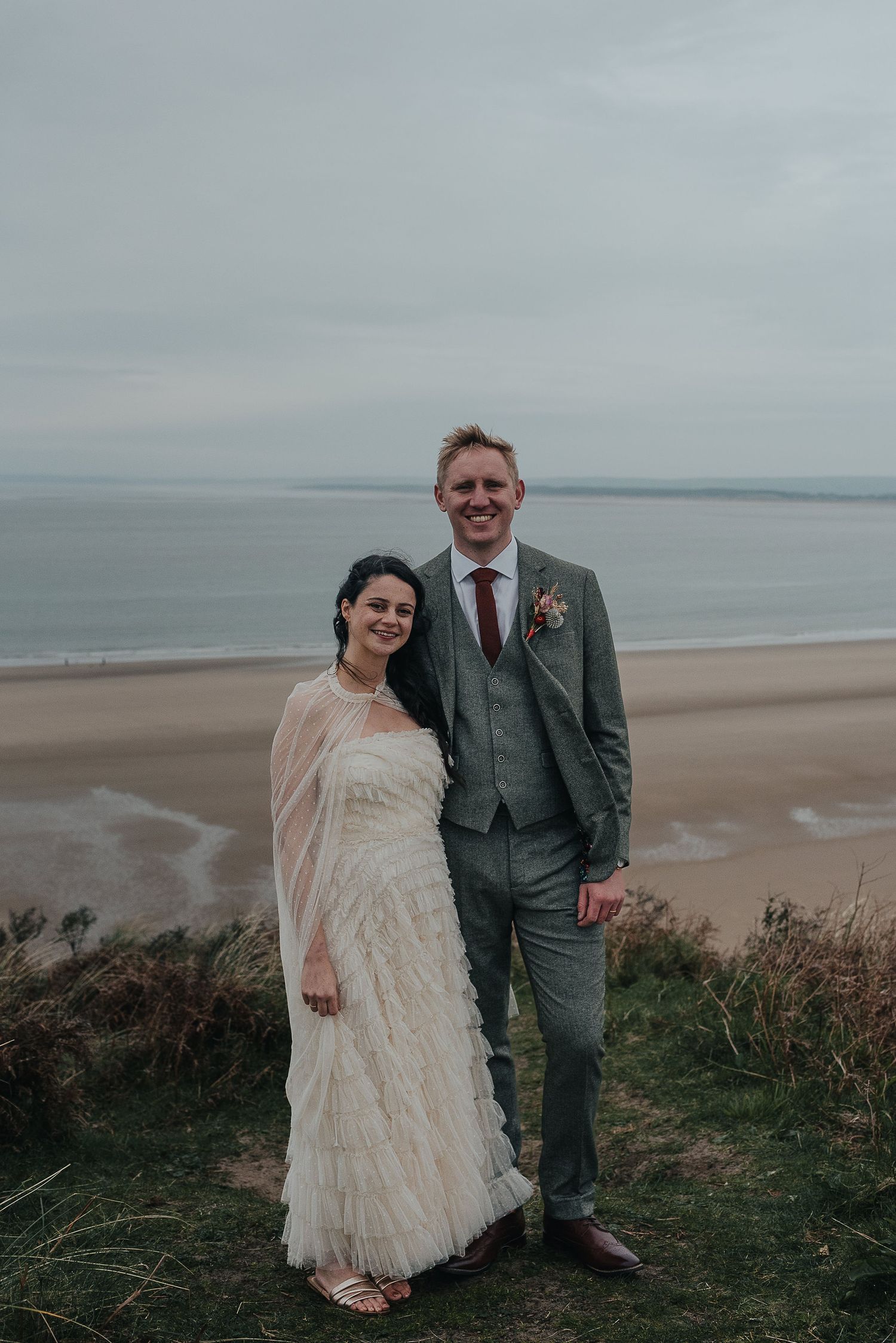 A couple poses for their wedding photos on a cliff overlooking a misty beach and ocean.