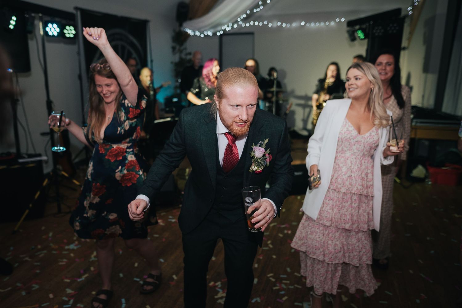 Wedding guests dancing and celebrating on dance floor with party lights in background.