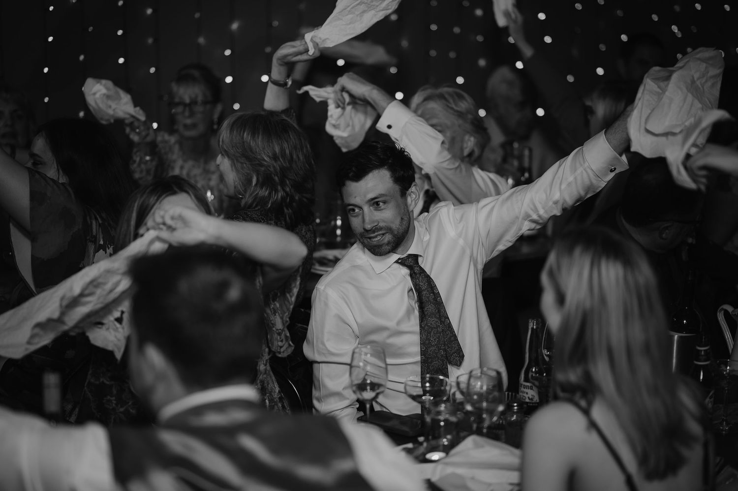 Wedding guests celebrate on dance floor with arms raised during reception party.