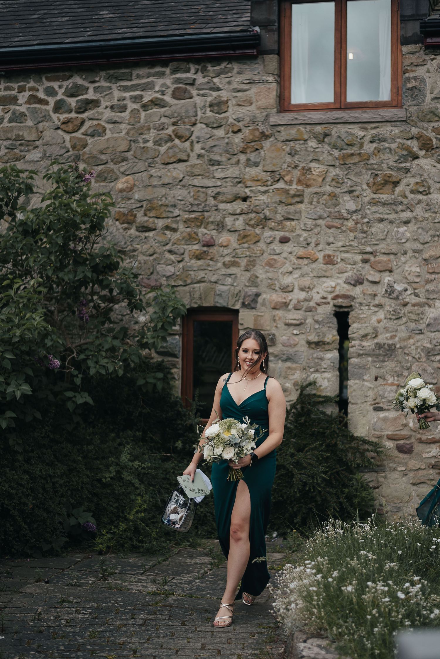 Navy dress-clad figure carrying a bouquet in front of a rustic stone wall with a window.