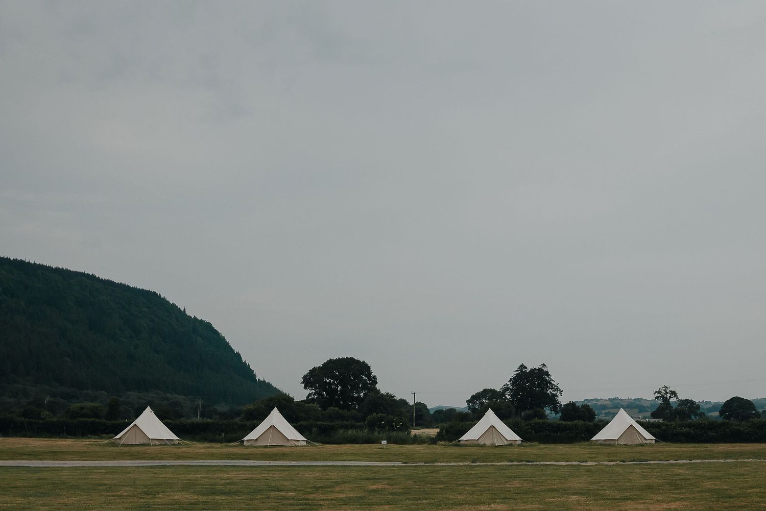 Four white bell tents set up in a field with mountains in the background under cloudy skies.