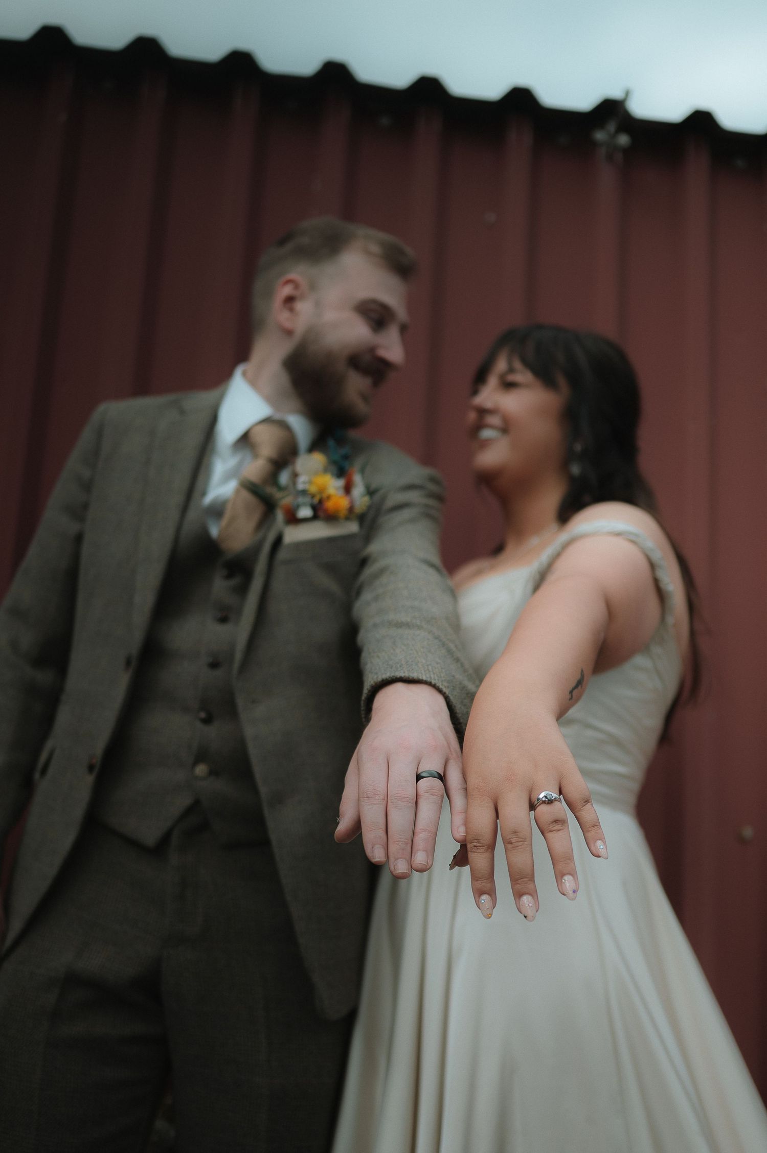 A couple in wedding attire poses together showing off rings against a rustic red barn backdrop.