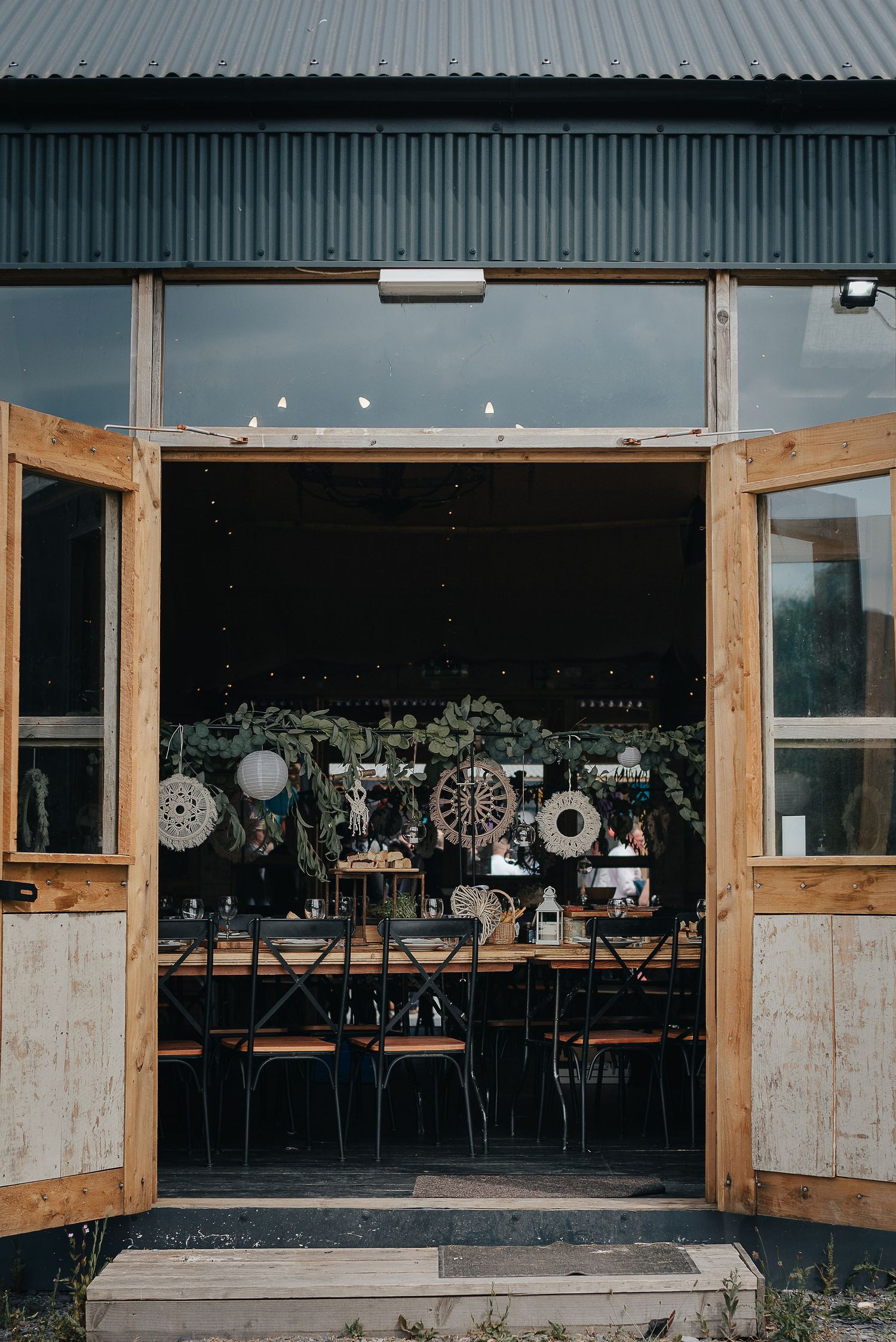 View through rustic wooden doors into a dark reception venue with hanging lights and decorative elements.
