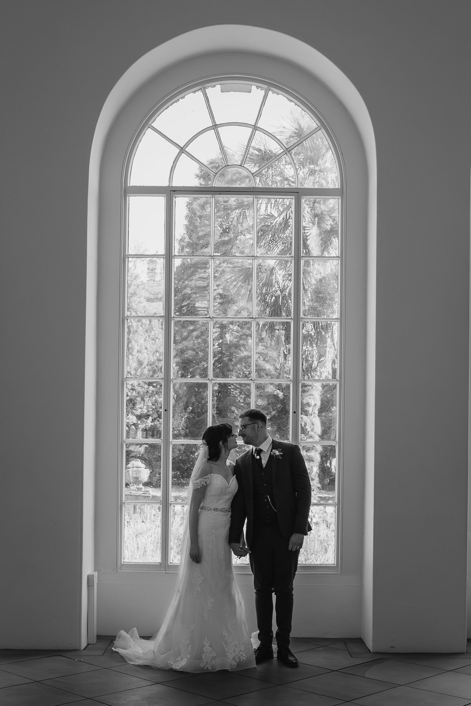Wedding couple silhouetted against large arched window in elegant indoor setting.