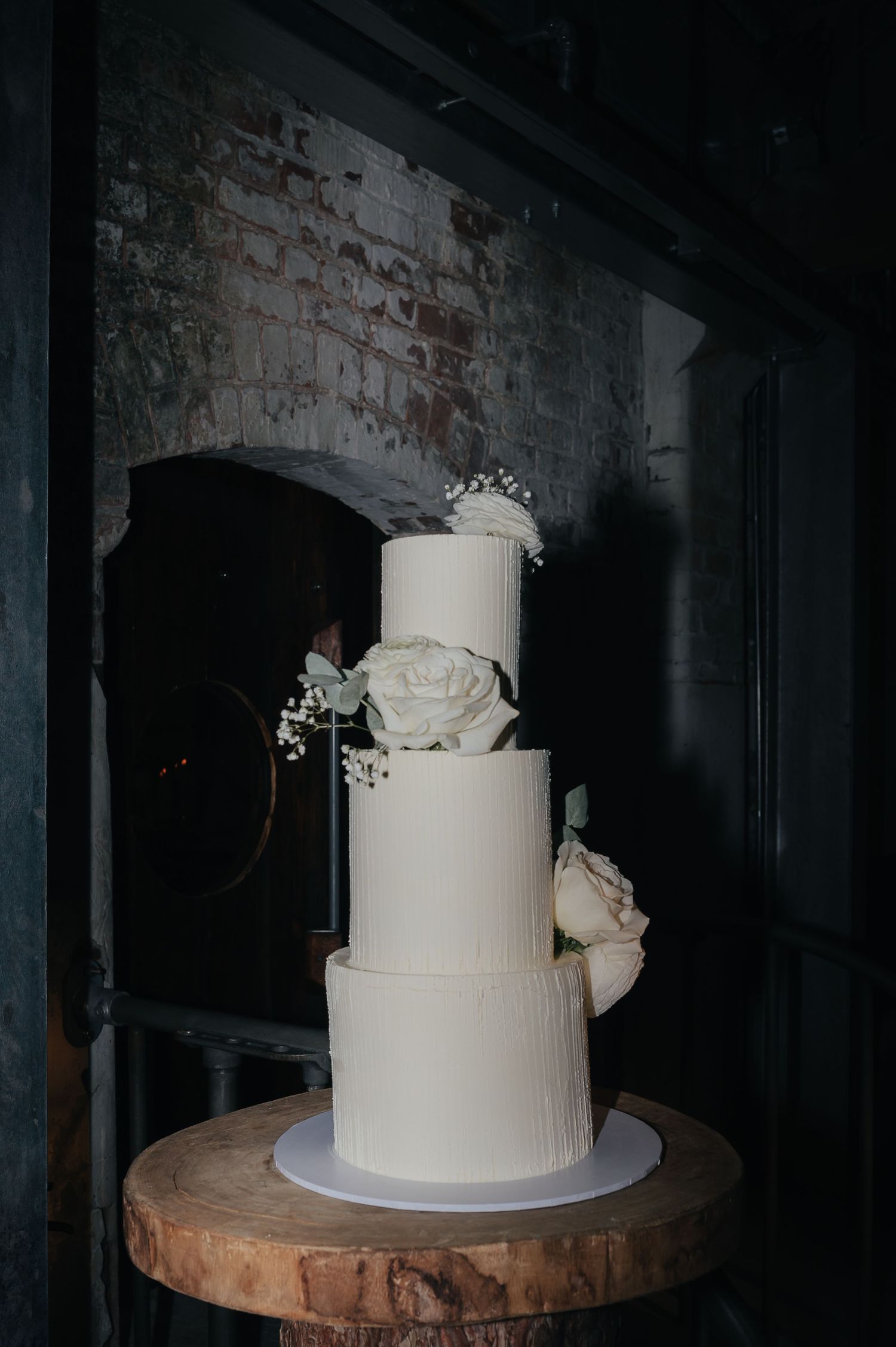 Elegant three-tier white wedding cake decorated with roses on a wooden stand against a brick wall.