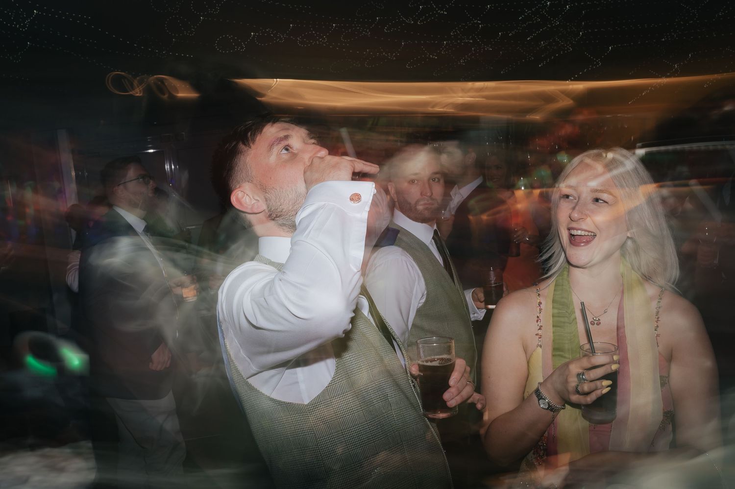 Party guests enjoying drinks and dancing in a dimly lit venue with motion blur effects.
