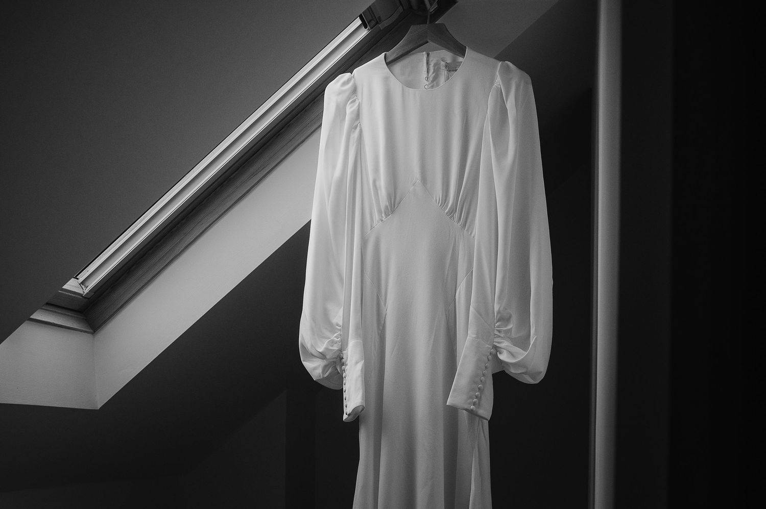 Elegant white wedding dress with long sleeves hanging under a skylight window.