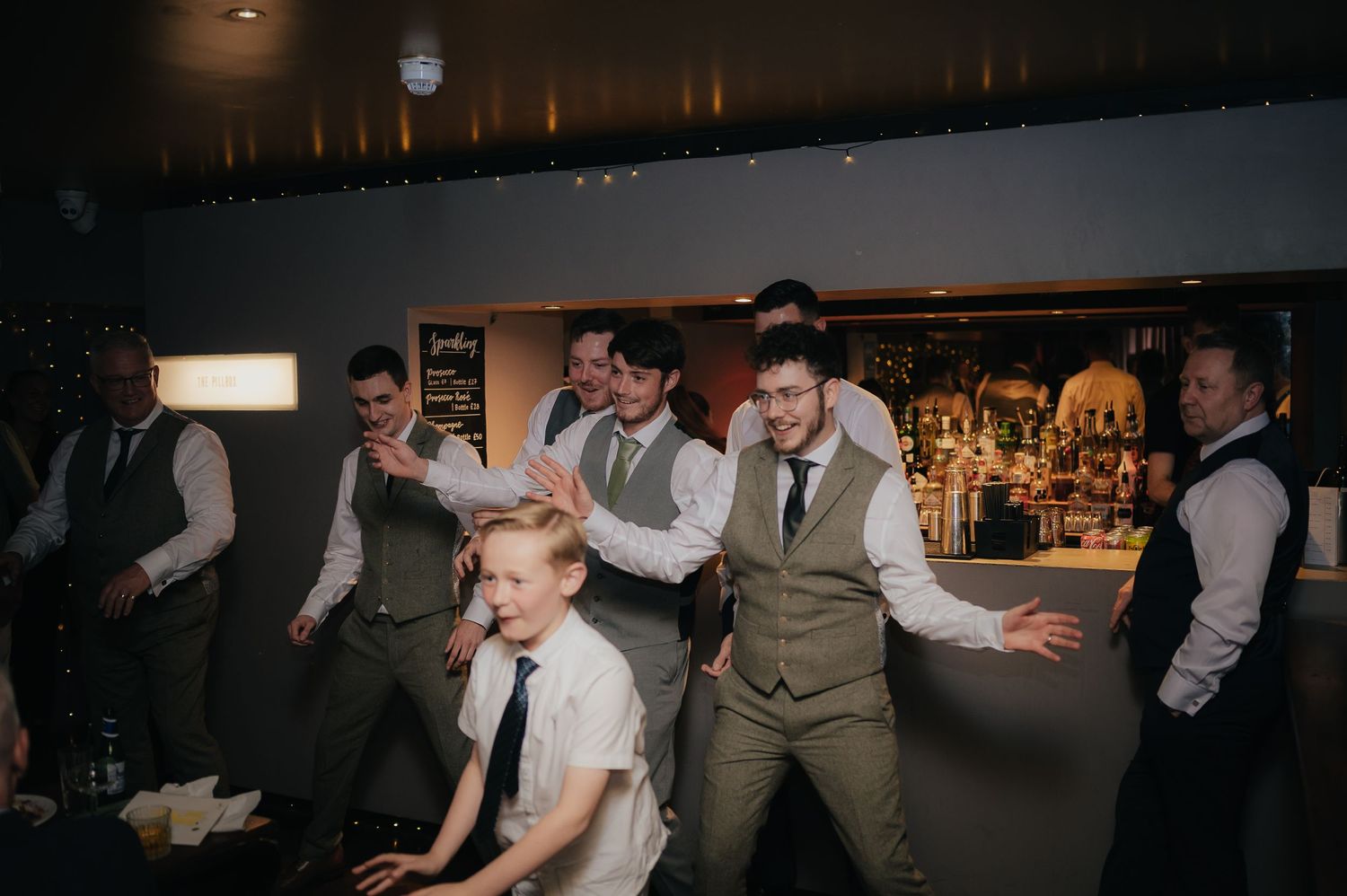 Groomsmen in matching gray vests strike playful poses at a wedding reception.