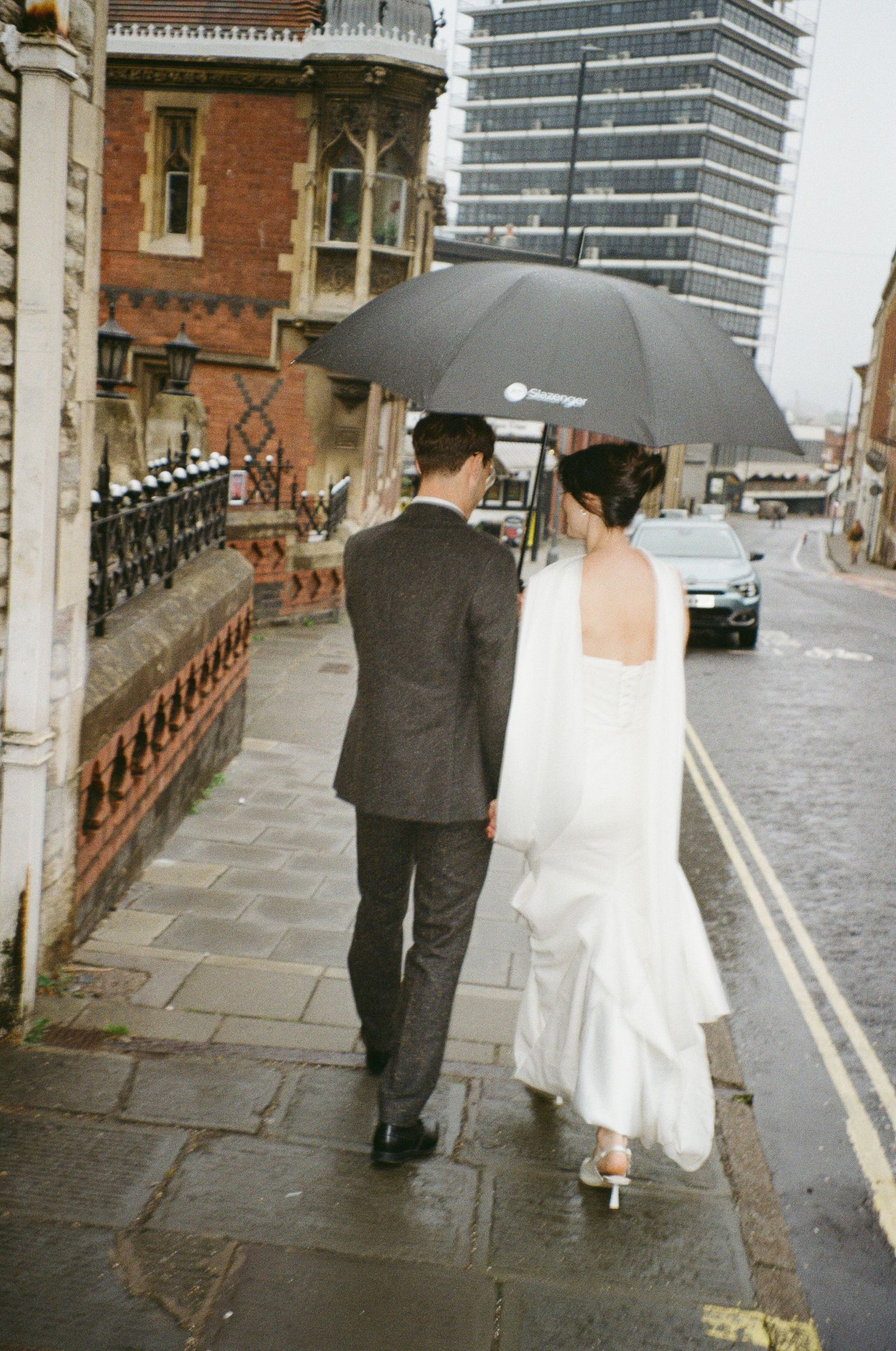 Bride and groom walking under black umbrella on rainy city street, bride in backless white gown, groom in grey suit.