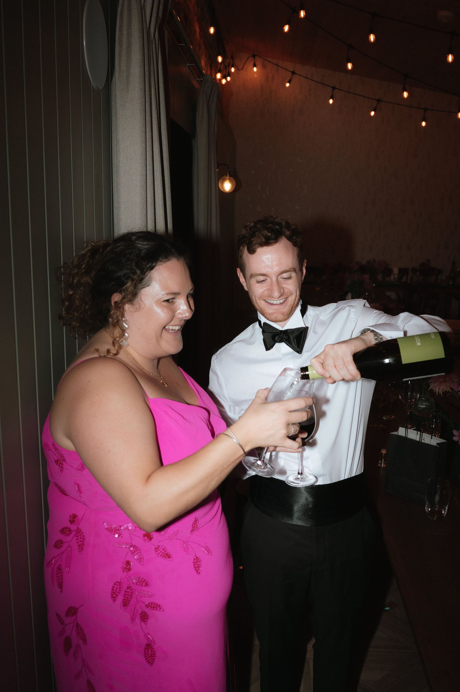 A guest in pink and server pour wine together at a wedding reception under string lights.