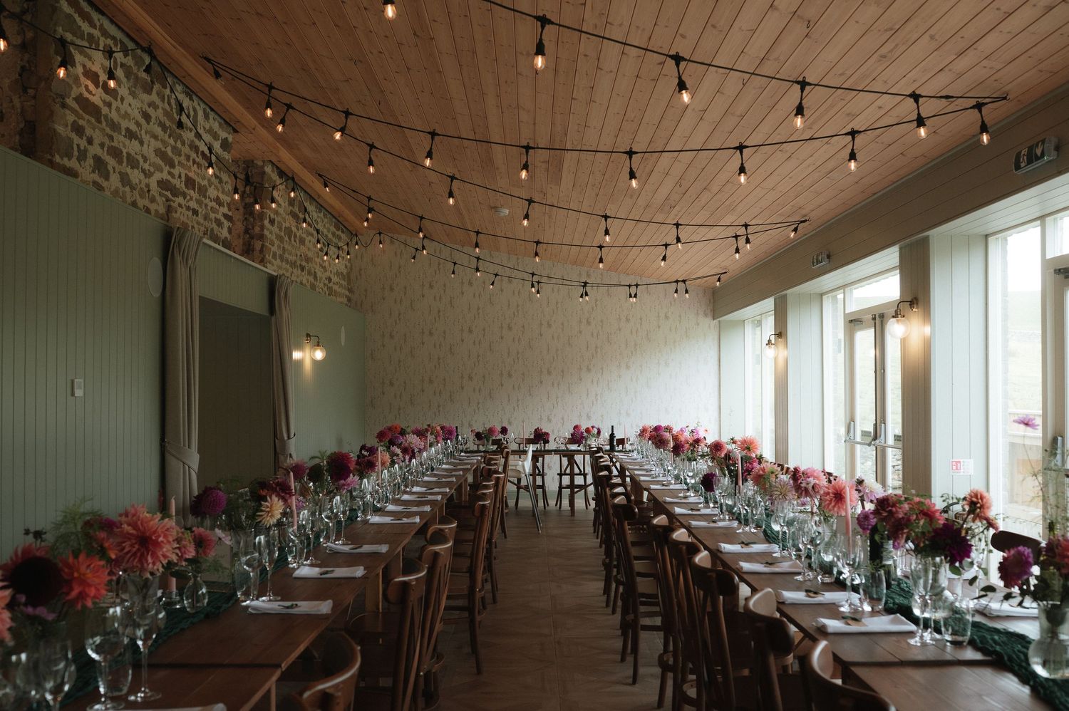 A long banquet hall with wooden ceiling features elegant table settings and pink floral arrangements under string lights.