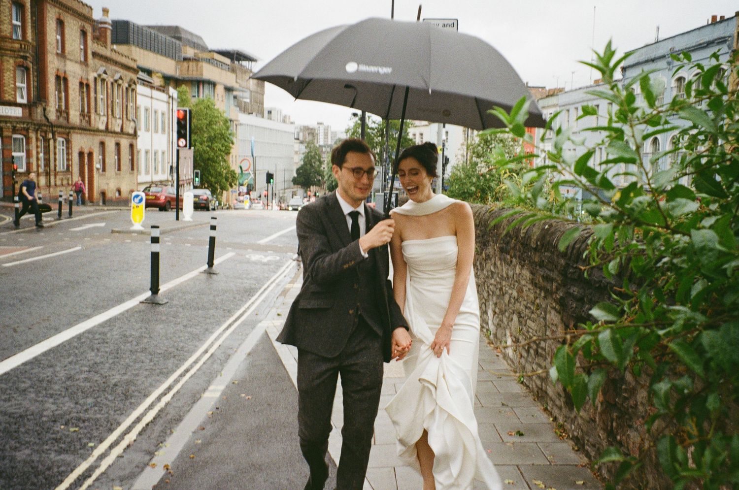 Bride and groom walking under a black umbrella on a rainy city street, bride in white strapless gown.