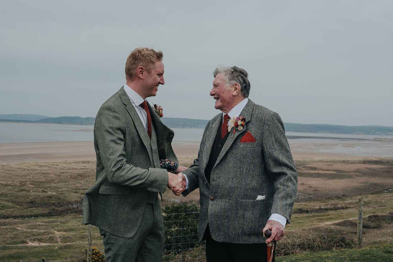 Two men in tweed jackets shake hands on a coastal clifftop.