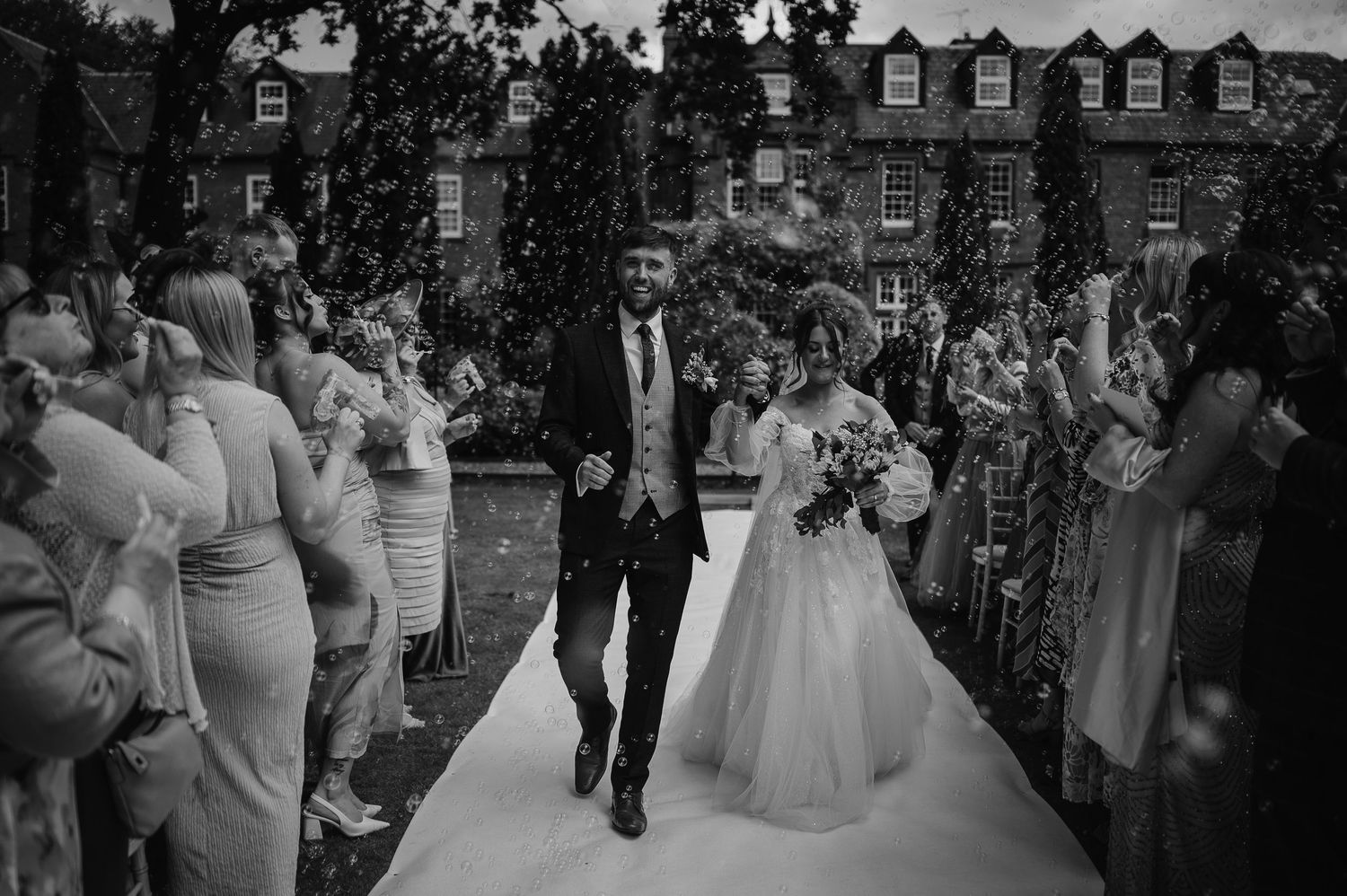 A black and white wedding photo of a couple walking down an outdoor aisle while guests celebrate.