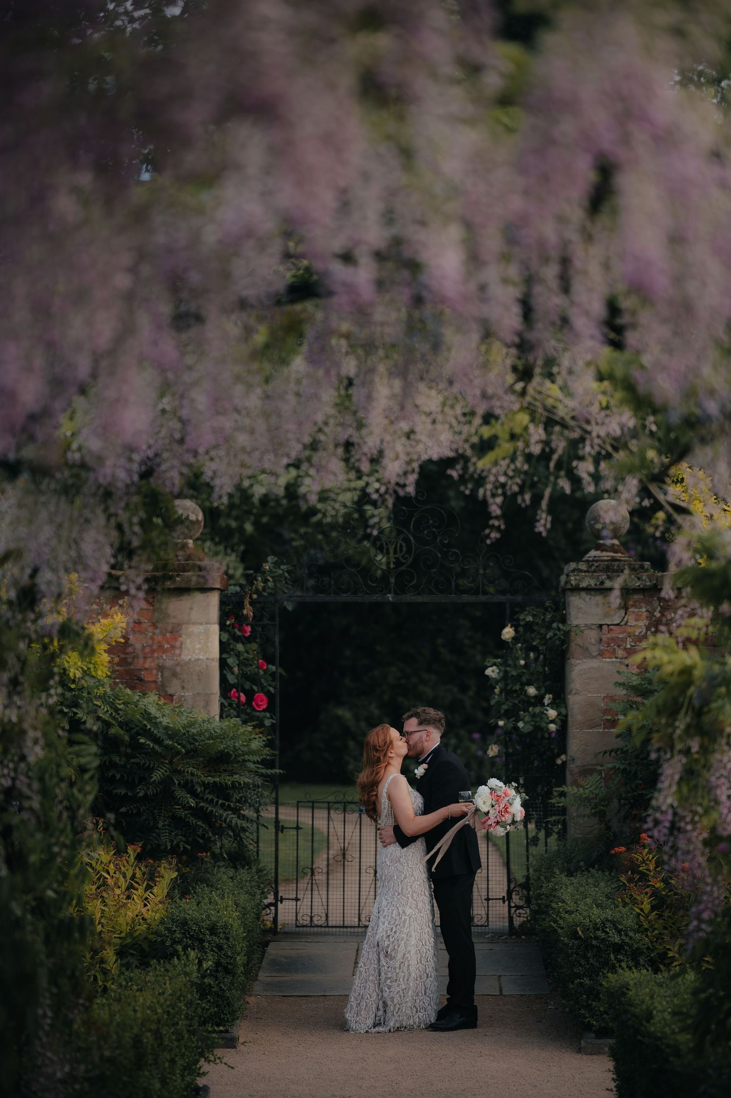 A romantic couple shares a dance moment at dusk in a garden setting framed by stone pillars and greenery.