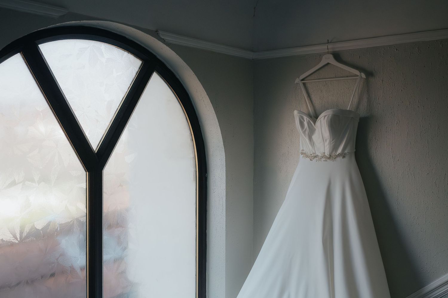 Elegant white wedding dress hanging by arched window in moody lighting.