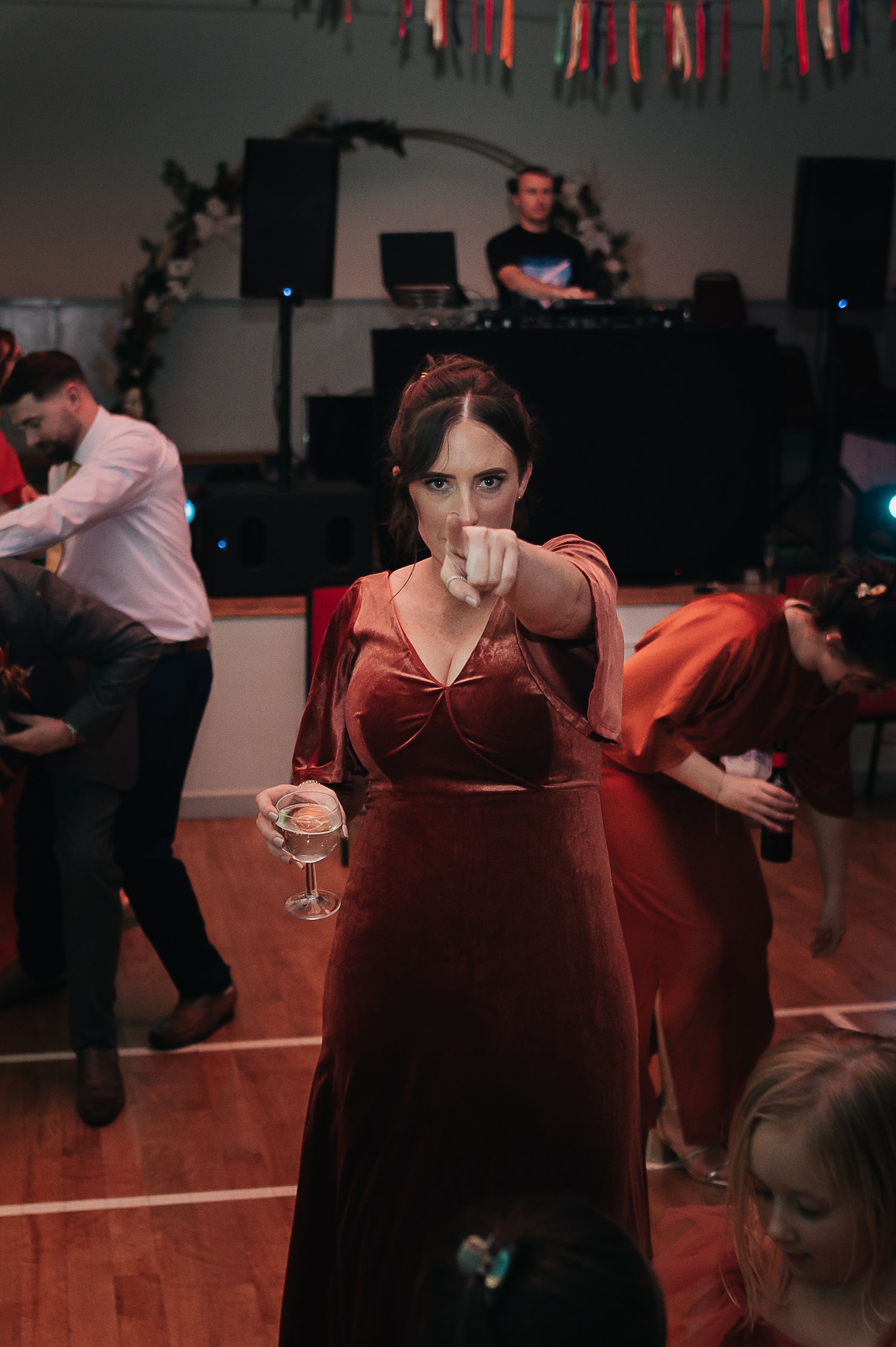 Woman in burgundy dress dances at wedding reception under string lights with DJ booth visible.