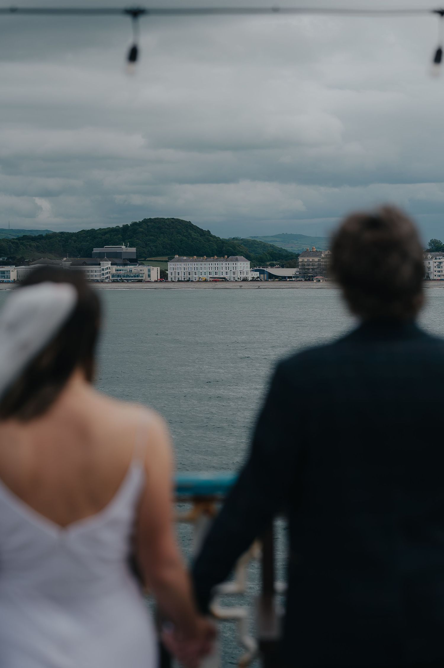 Couple overlooking a coastal bay view with mountains and buildings in the distance.