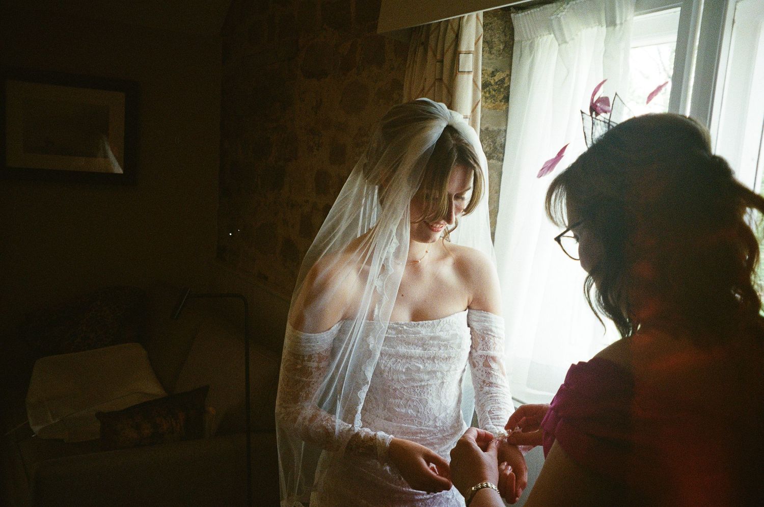 Bride in lace wedding dress and veil being helped by bridesmaid near window in soft natural light.