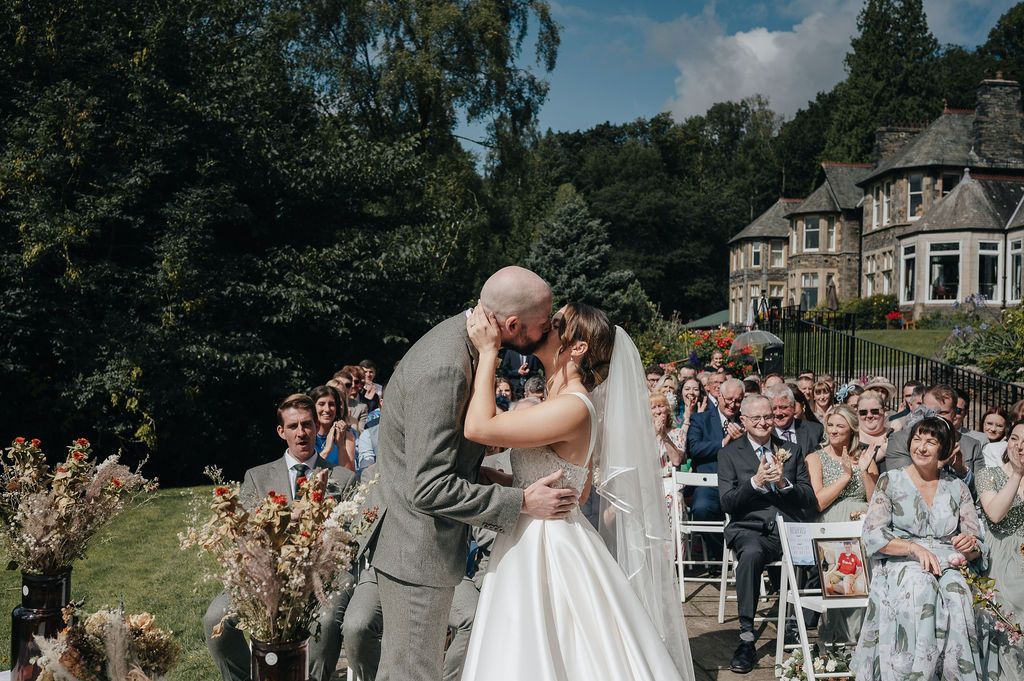 Newlyweds sharing their first kiss during outdoor ceremony with guests looking on.