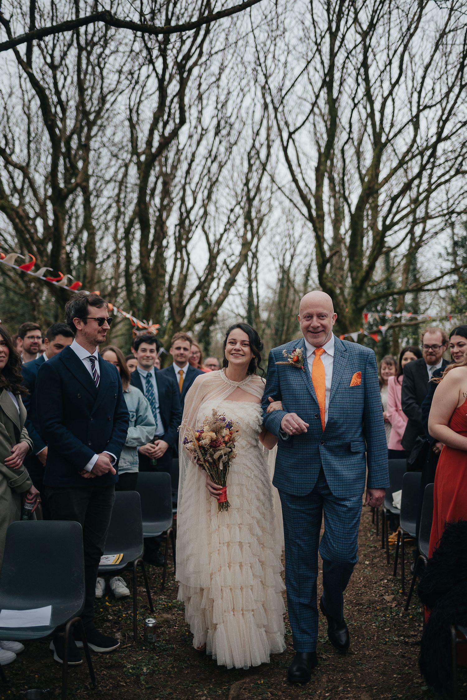 Wedding ceremony procession along tree-lined path with seated guests.
