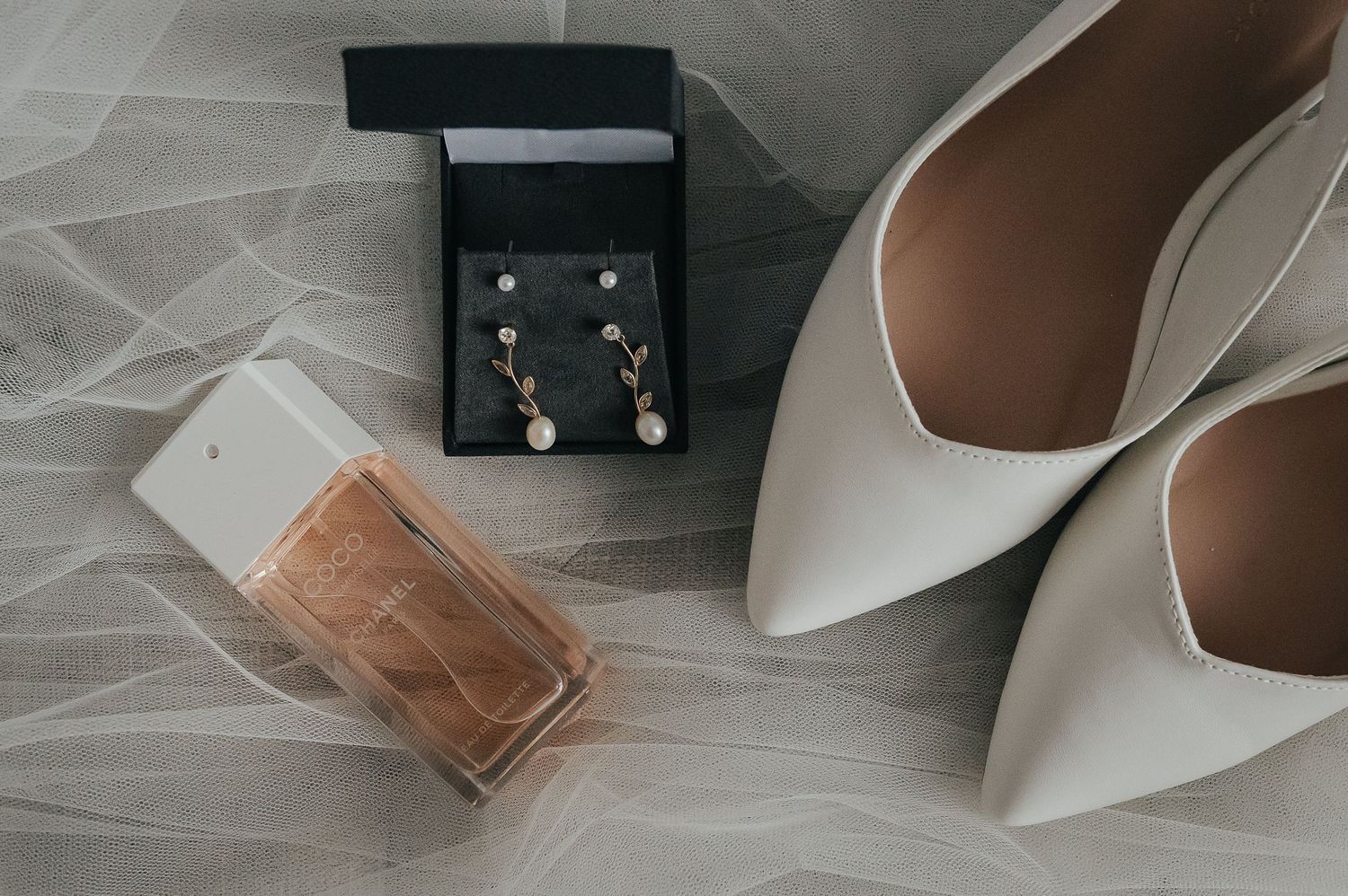 Wedding accessories including white heels, pearl earrings and perfume bottle arranged on lace.